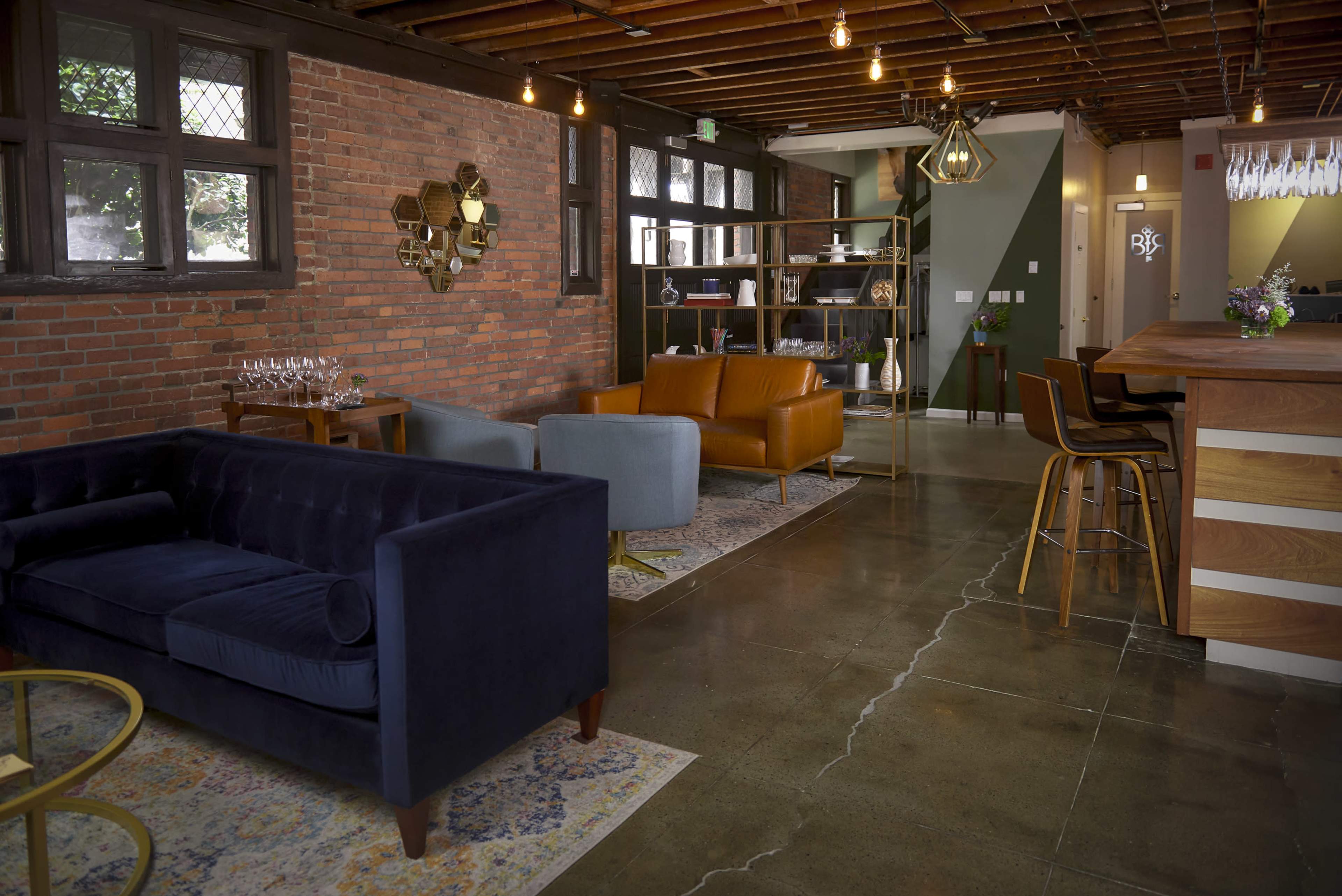 The image shows a modern lounge area featuring a mix of blue and brown upholstered seating, wooden shelving, and pendant lighting against a backdrop of exposed brick walls.