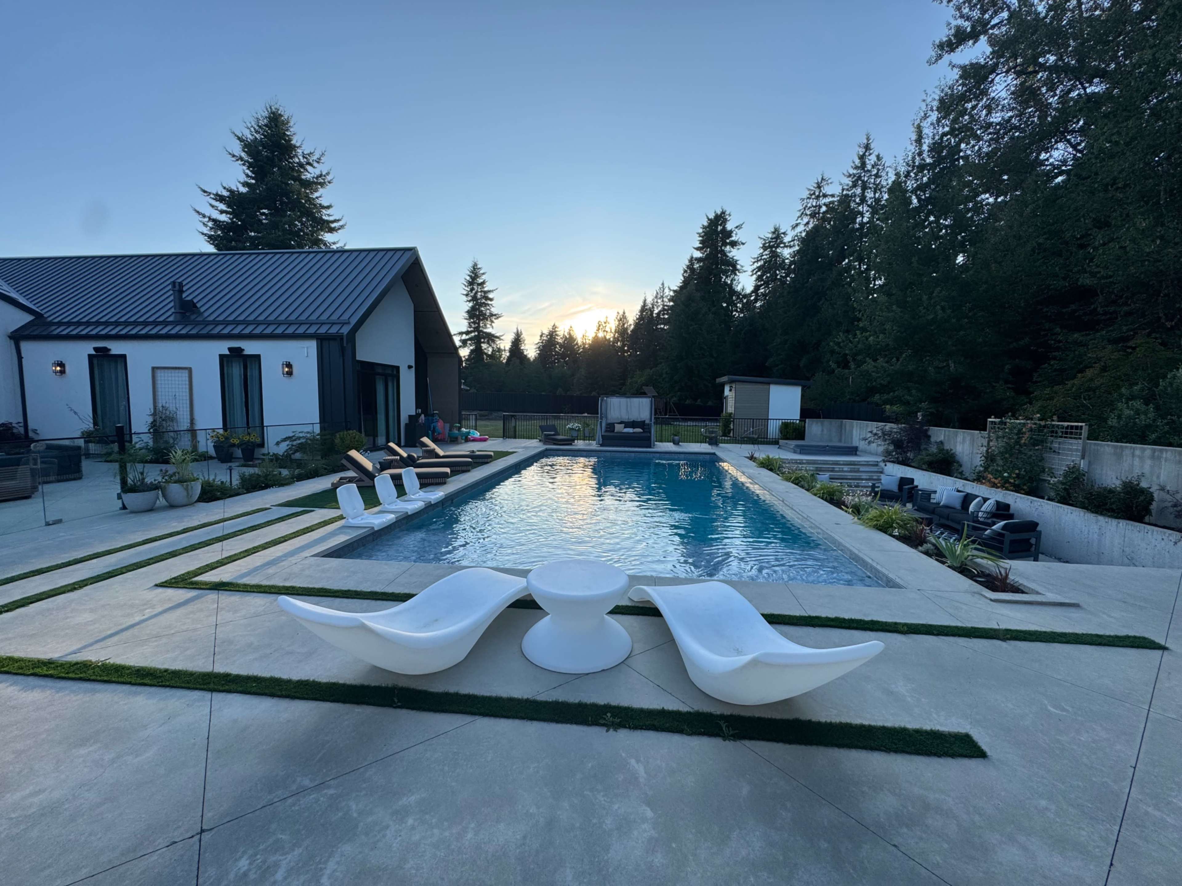 The scene features a modern pool area with two white lounge chairs positioned at the water's edge, surrounded by landscaped greenery and a contemporary house in the background.