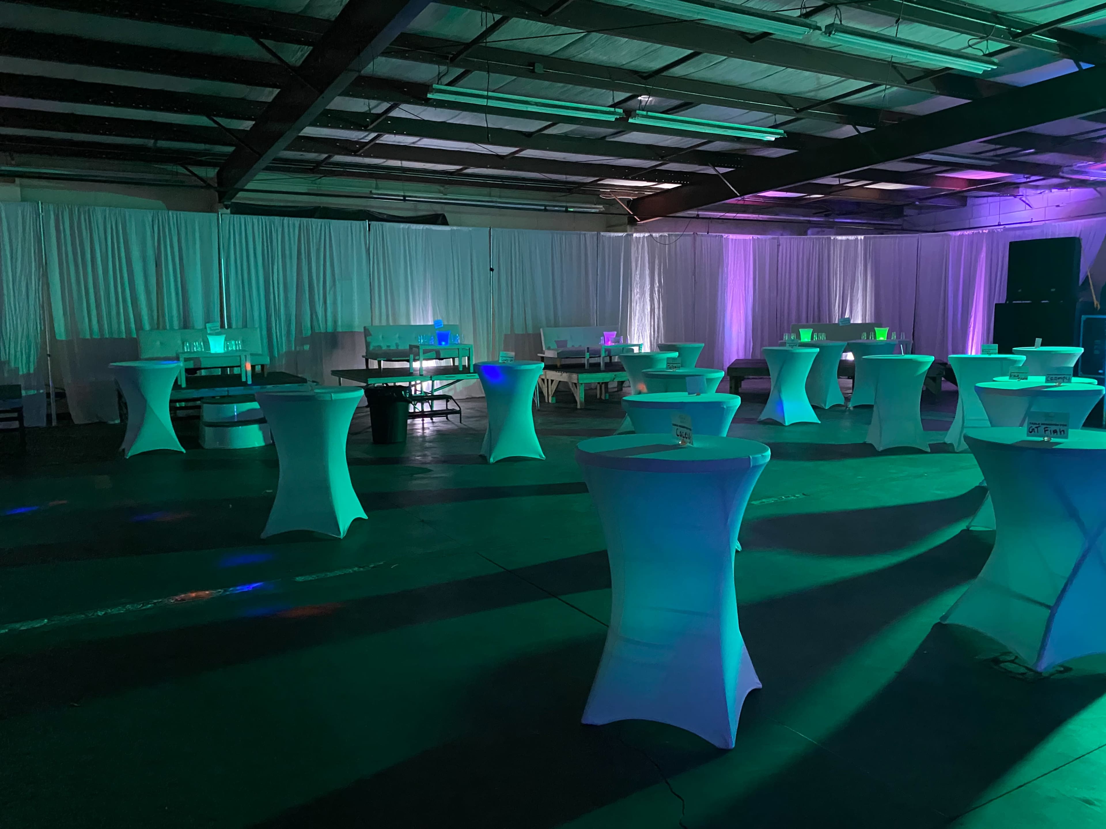The image shows an event space with illuminated tables arranged throughout, surrounded by white drapes and varying colored lights.