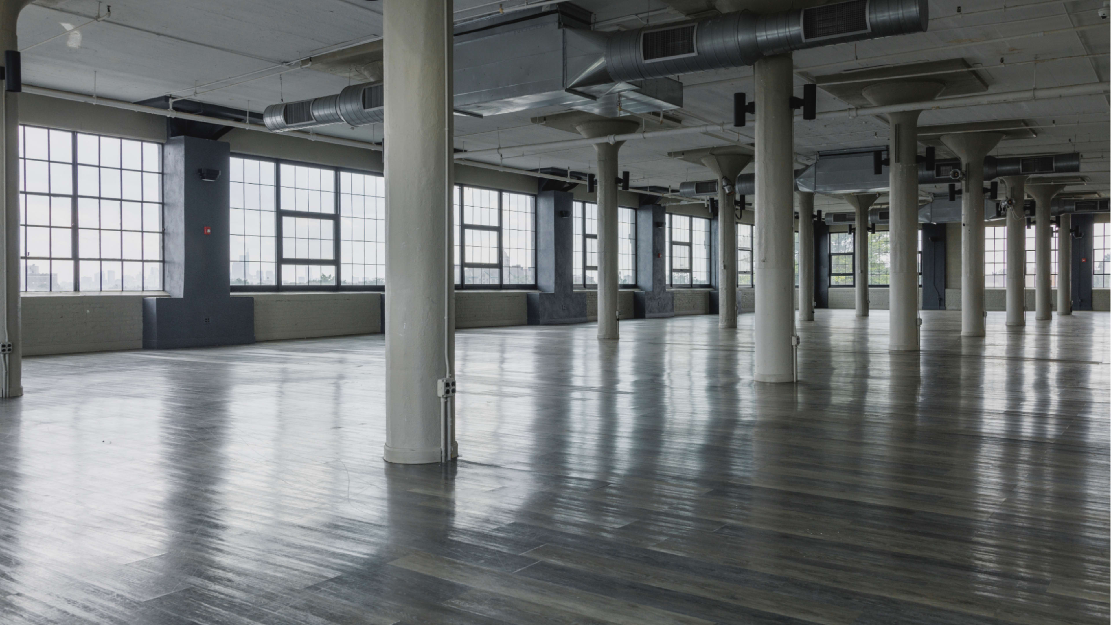The image shows a spacious, empty industrial-style interior with large windows and polished wooden floors, supported by concrete pillars.