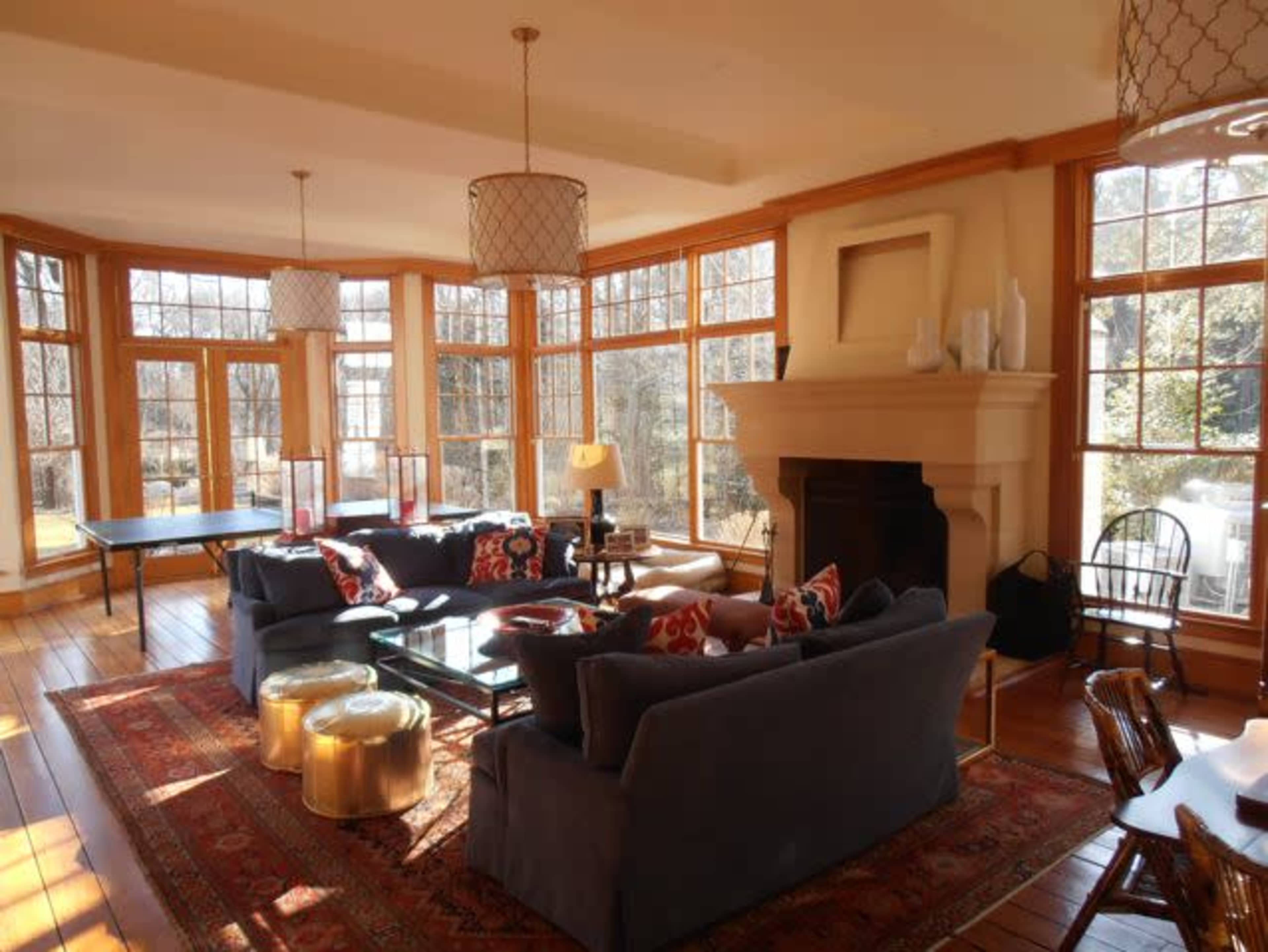 A bright living room features large windows, comfortable seating, and a central fireplace, with decorative tables and lamps.