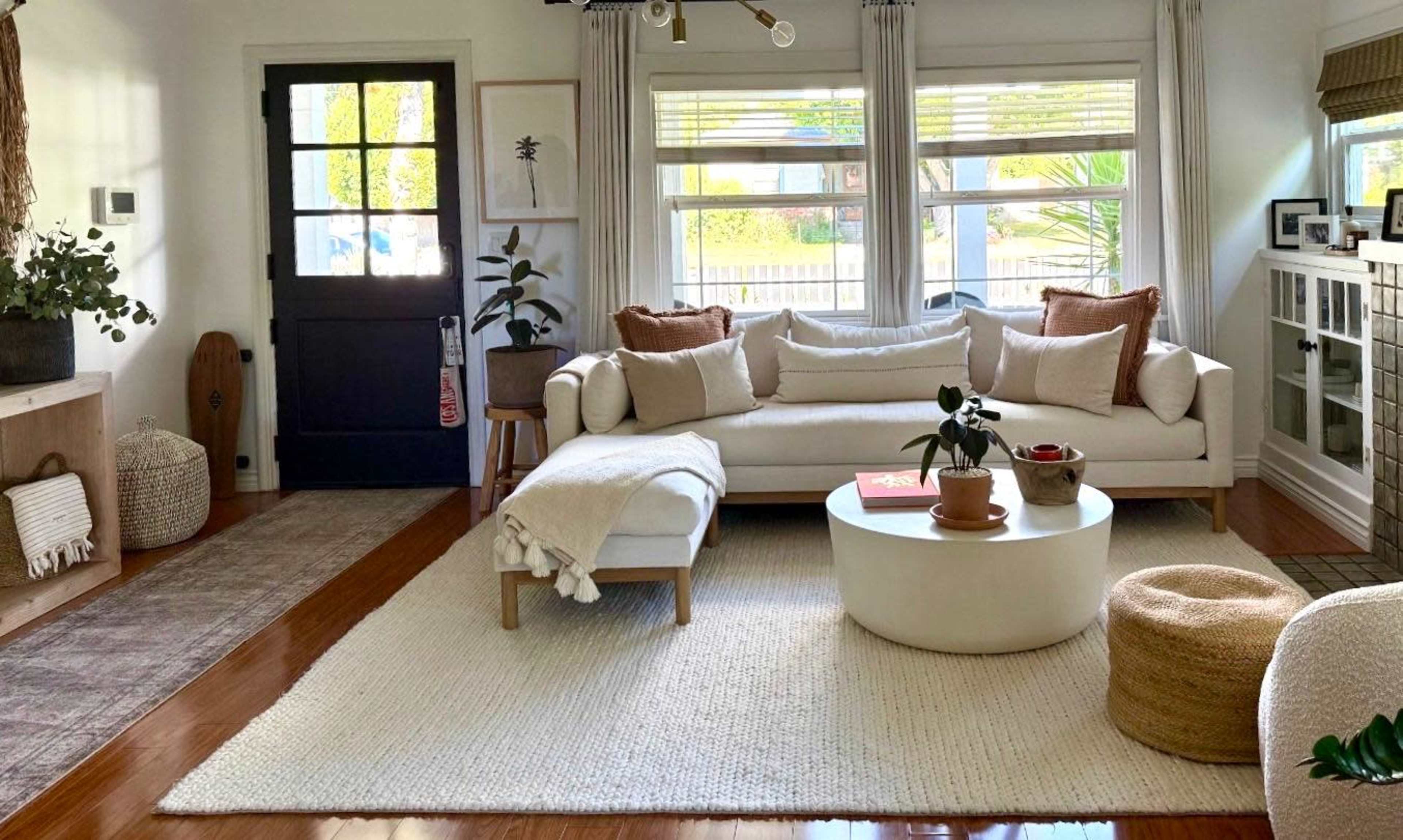The image shows a bright, modern living room with a white sectional sofa, a round coffee table, and natural decor elements like plants and woven baskets.