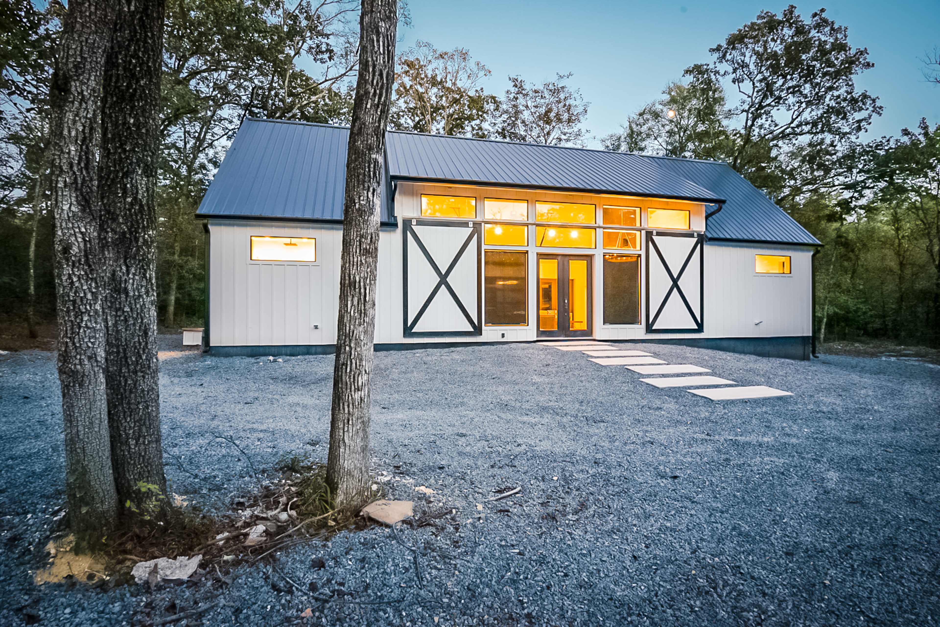 A modern, two-story house with a metal roof and large windows is set in a wooded area, featuring a gravel driveway leading to its entrance.