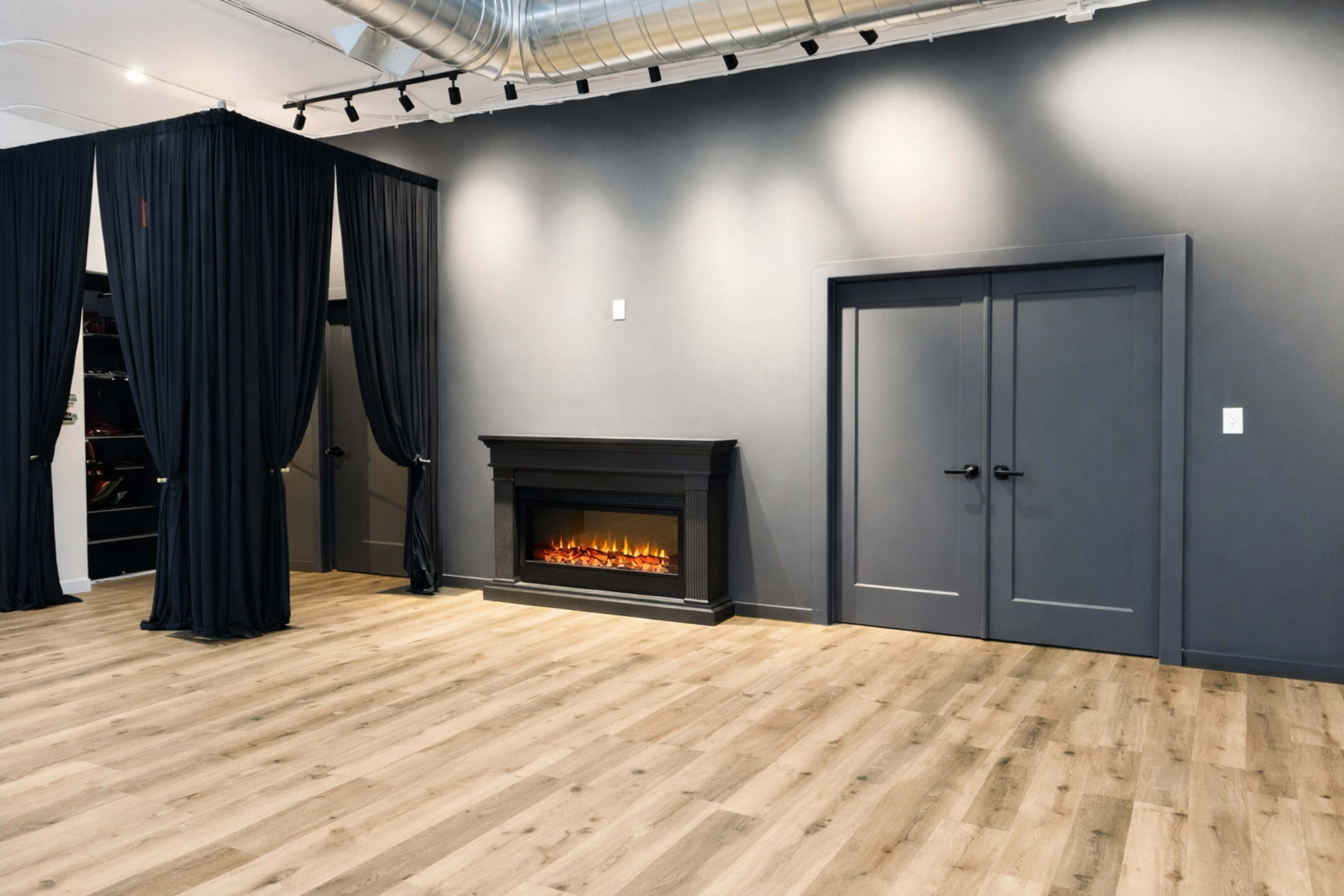 The image shows a spacious interior with a faux fireplace against a dark wall, flanked by two closed doors, and black curtains on the left side.