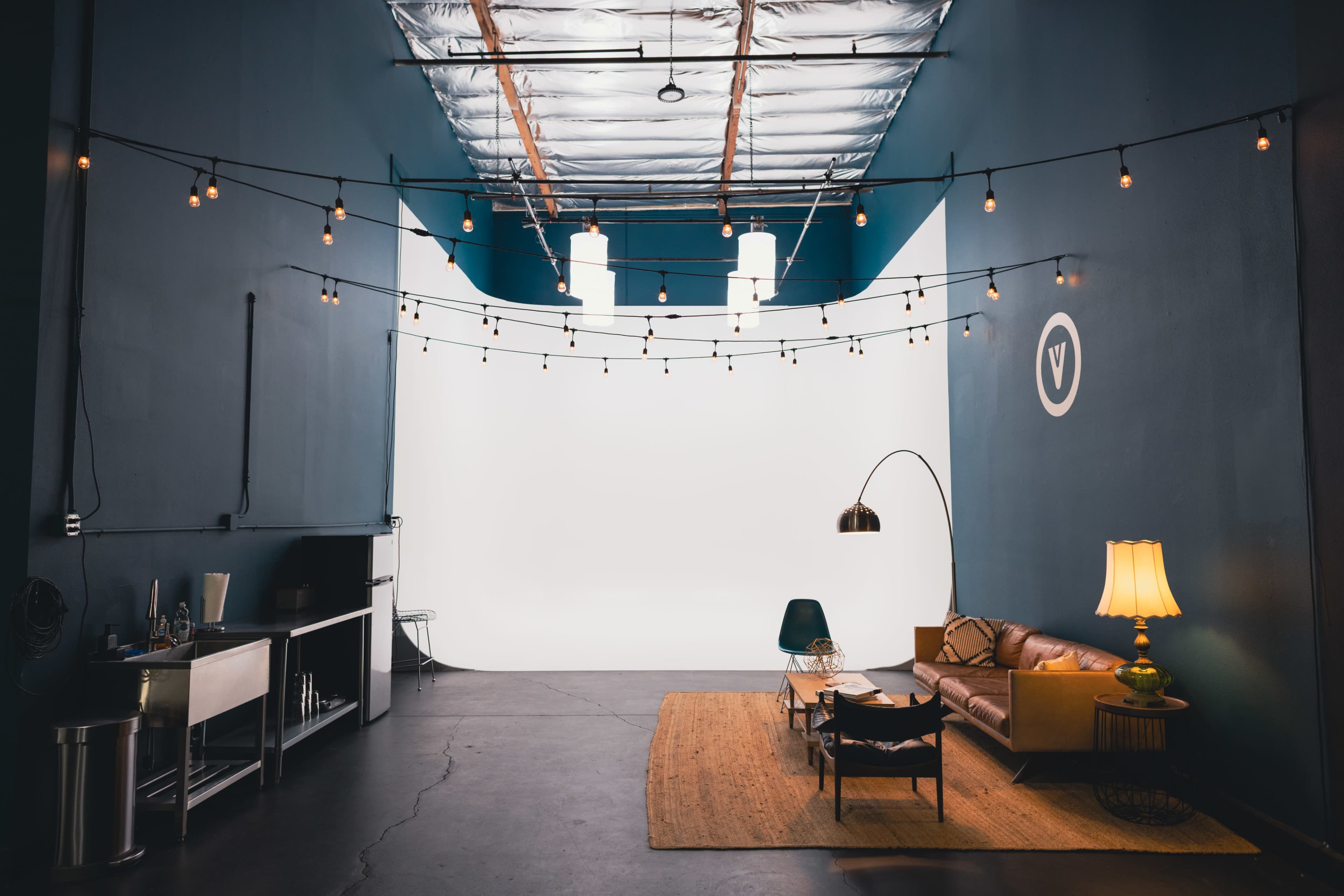 The image shows a spacious, industrial-style room with blue walls, string lights overhead, and a furnished sitting area featuring a sofa, lamp, and rug.