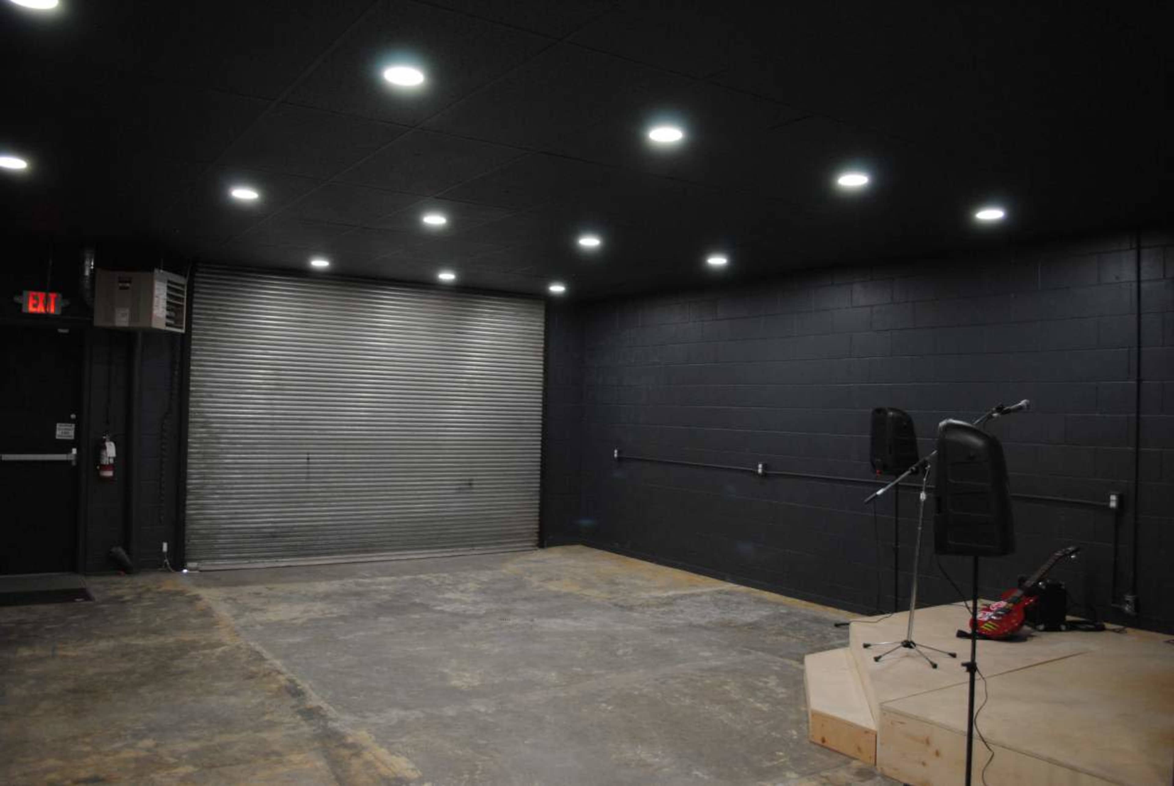 The image shows a dimly lit room with black walls, a large metal roll-up door, a wooden stage with a microphone stand, and an electric guitar resting on the floor.