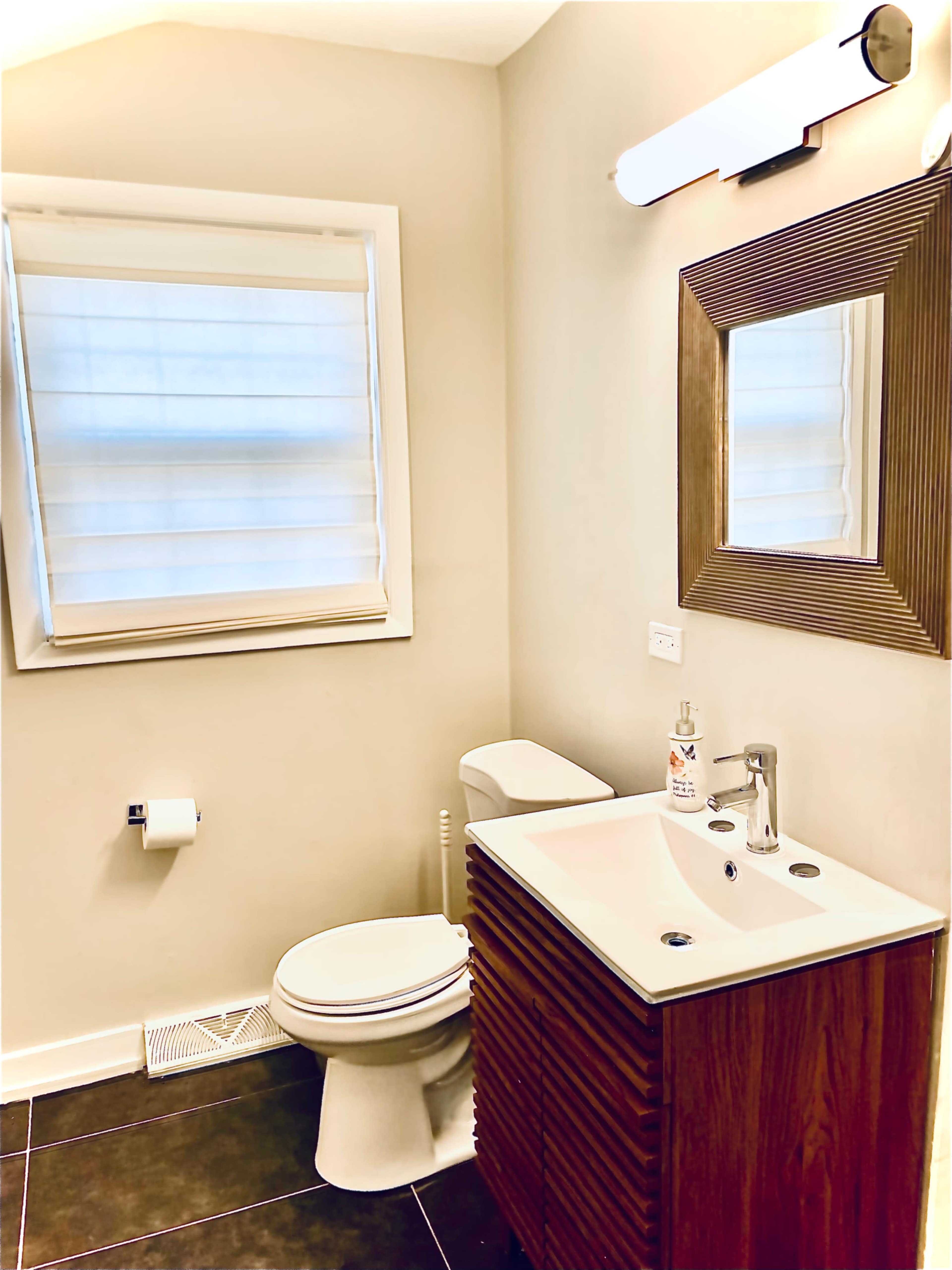 A small bathroom with a toilet, a sink with a wooden vanity, a mirror, and a window covered by a light-colored shade.