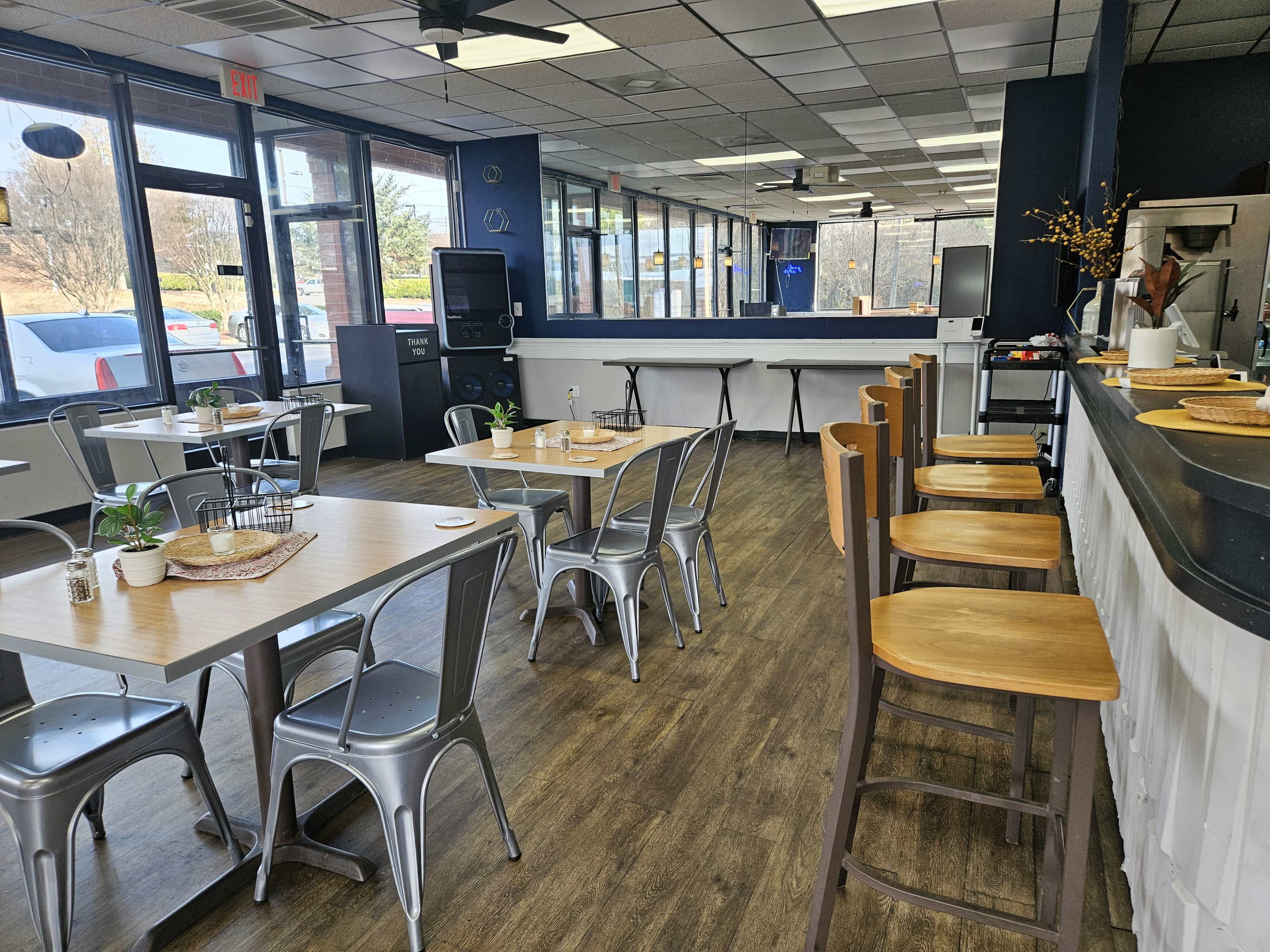 The image shows a spacious, modern café interior with several tables, chairs, and a counter, featuring large windows that allow natural light to enter.