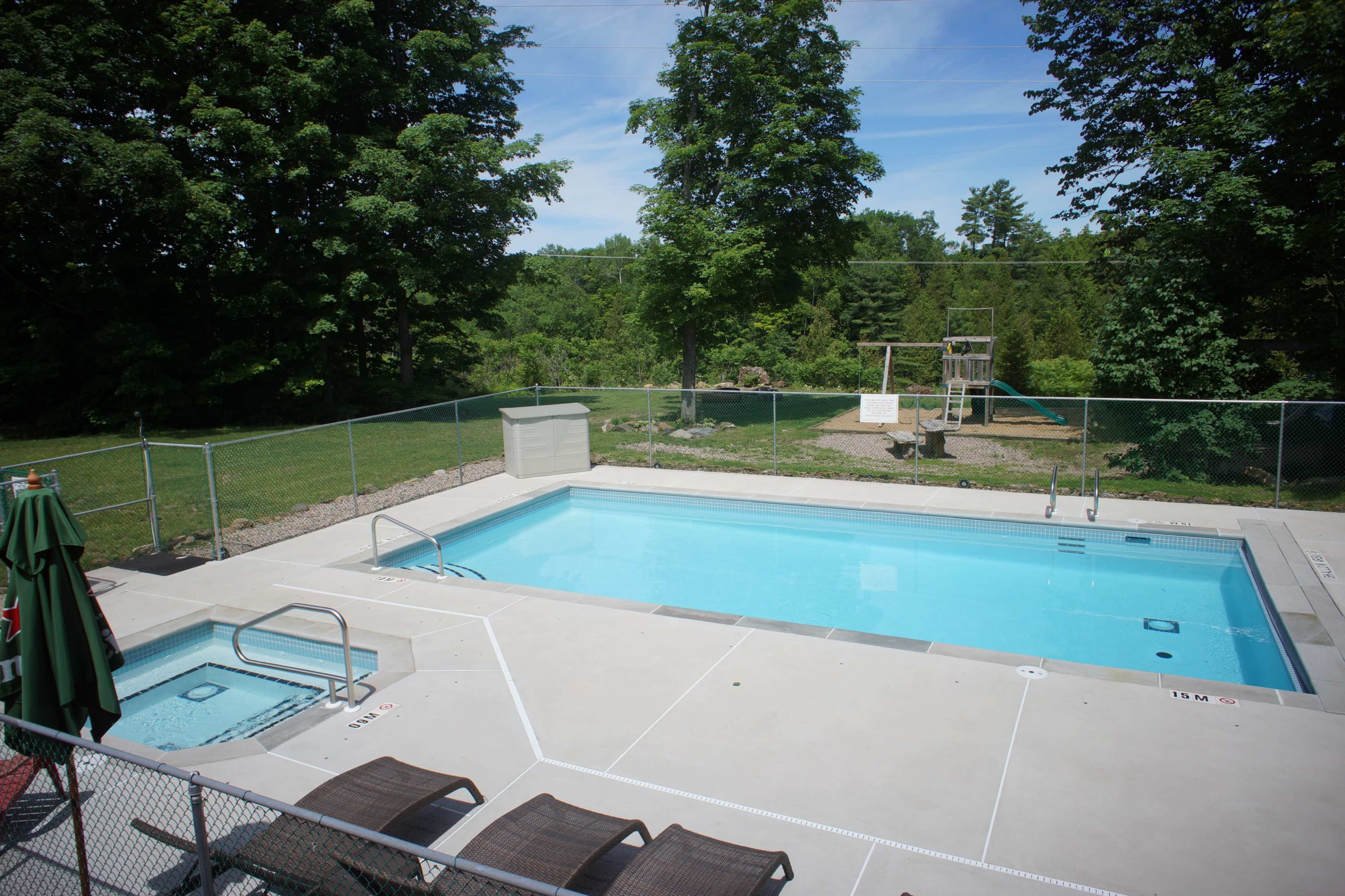 The image shows a rectangular swimming pool surrounded by a fenced area, with a hot tub and chairs nearby, and playground equipment in the background.