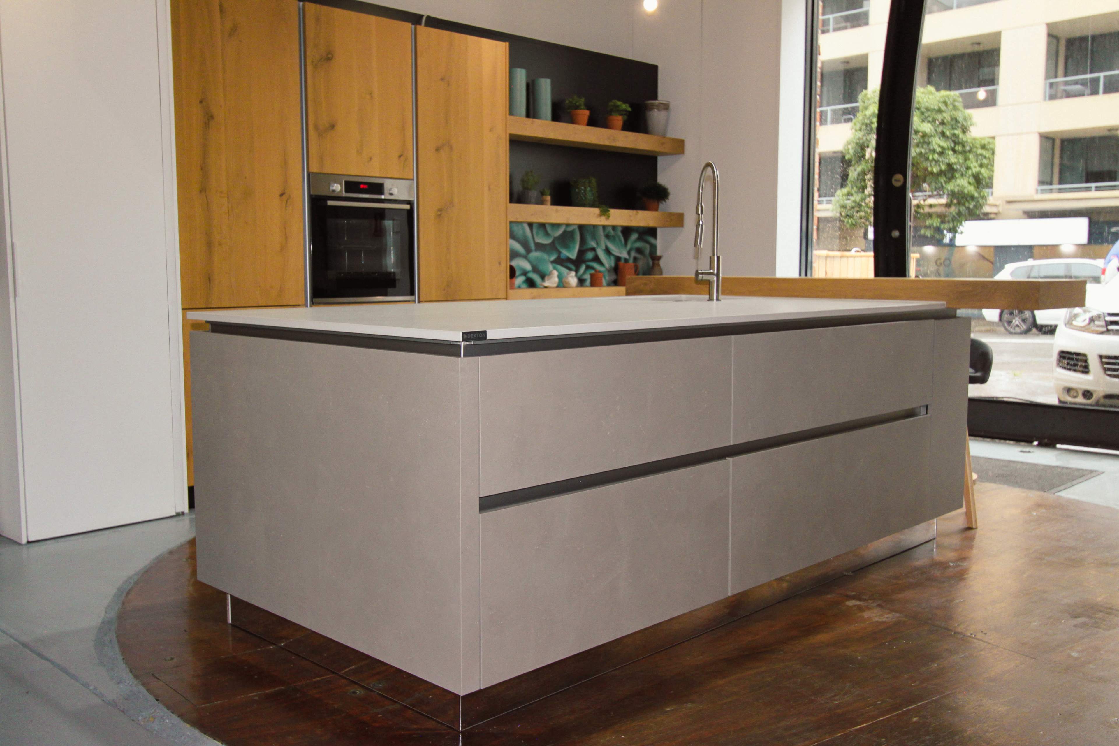 The image features a modern kitchen with a central island, gray concrete countertops, and wooden cabinetry with open shelving displaying plants.