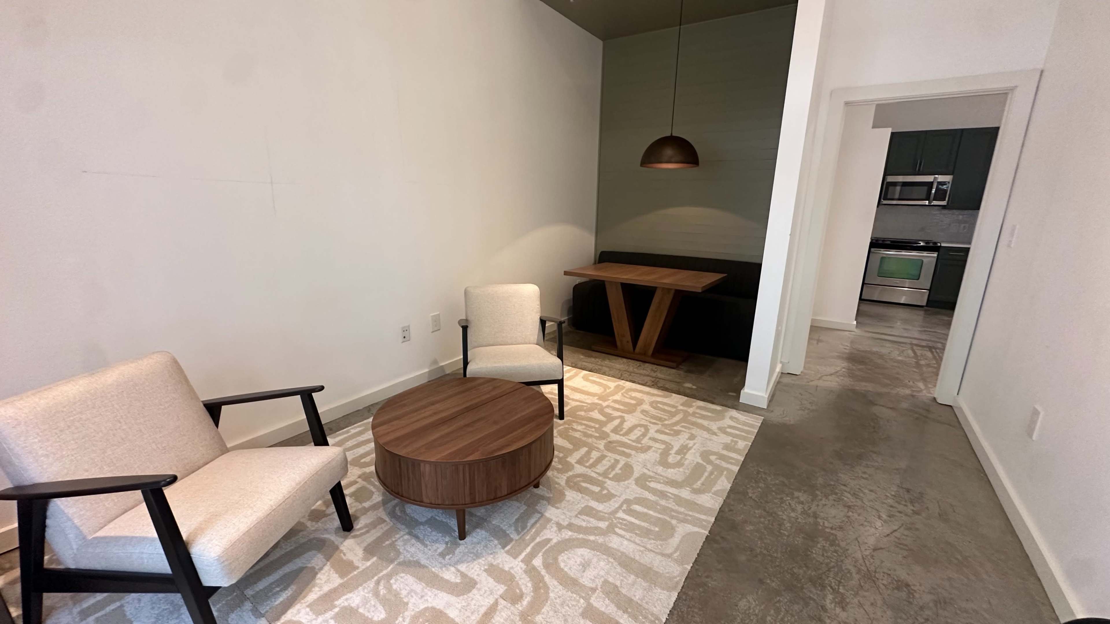 The image shows a minimalist interior space featuring two chairs, a round coffee table, and a dining area with a pendant light and a rectangular table.