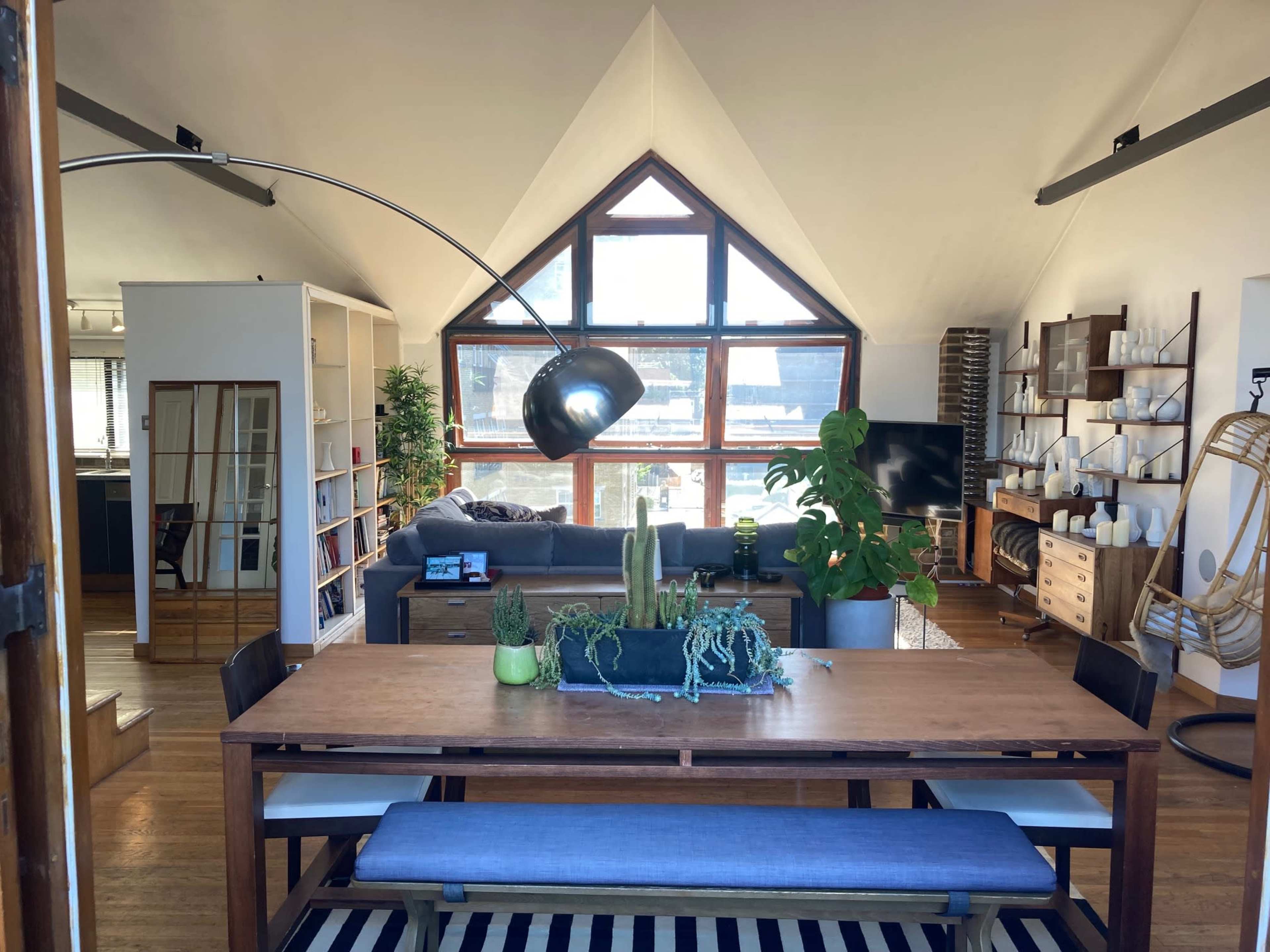 The image shows a modern, open-concept living space featuring a dining table at the foreground, a couch and television in the background, and large windows that provide natural light.