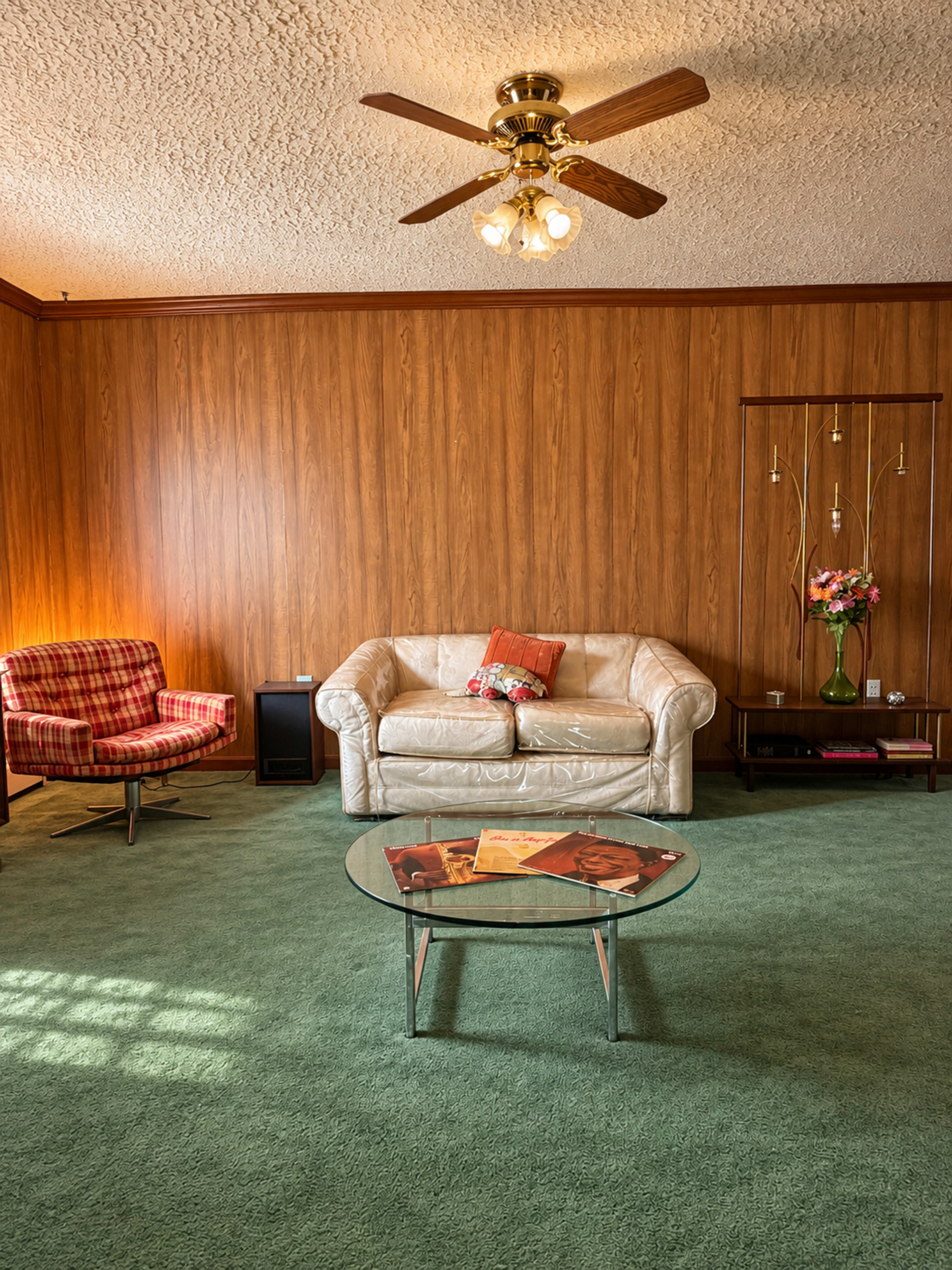 The room features a green carpet, a light-colored leather sofa, a checkered armchair, a glass coffee table, and wood-paneled walls with a ceiling fan.