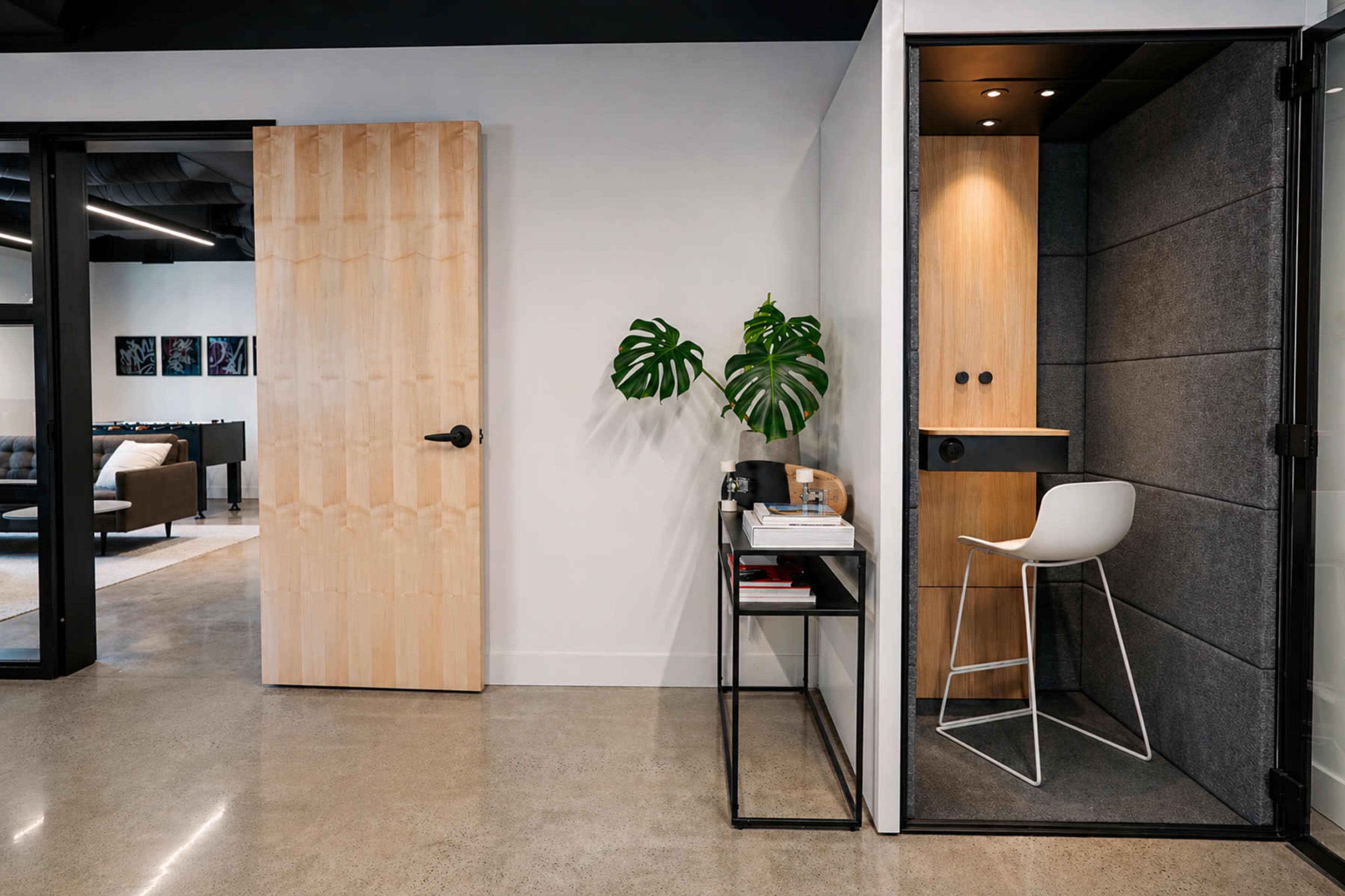 The image shows a modern office space featuring a wooden door, a green plant, a small desk with books, and a soundproof booth with a stool.