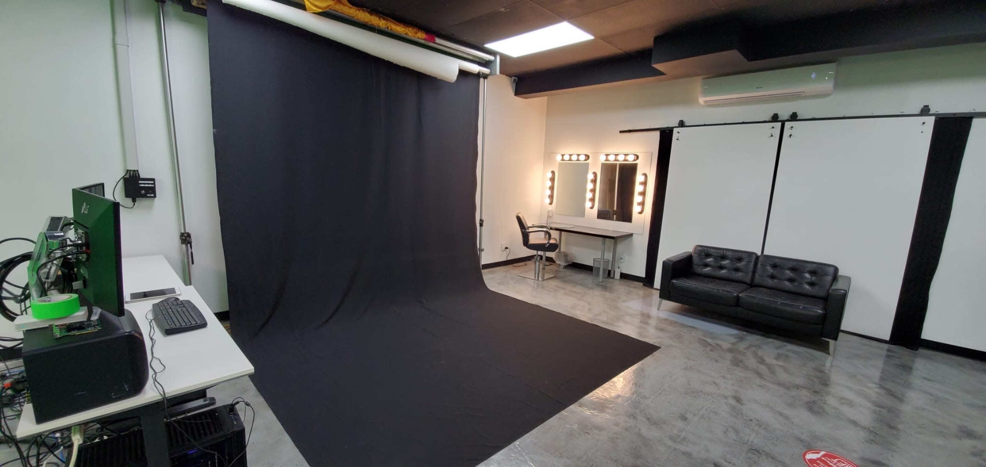 The image shows a photography studio with a black backdrop, a makeup station with lights, a black couch, and a workstation with computers.