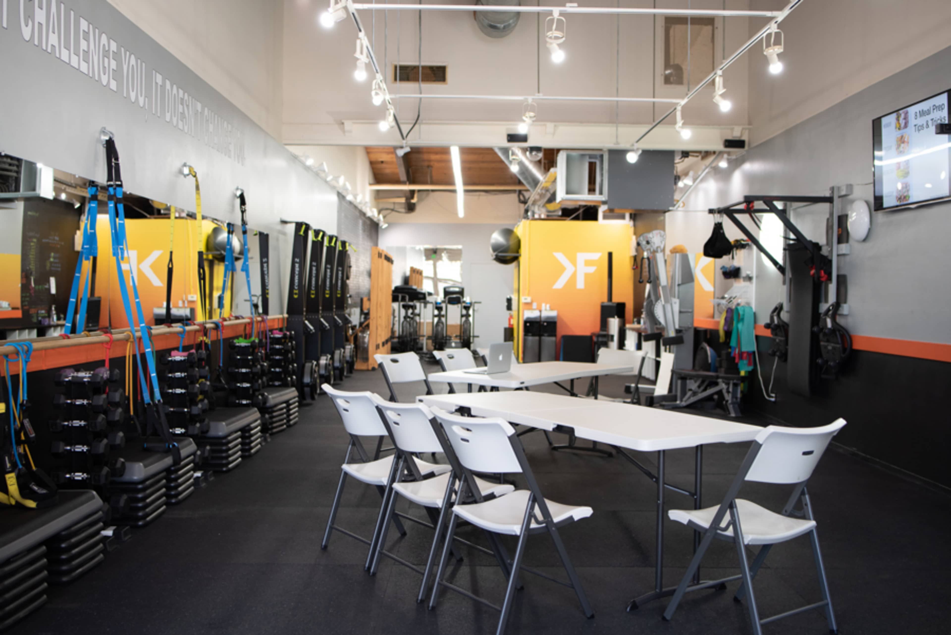 The image shows a fitness studio featuring workout equipment, mirrors, and a small seating area with white chairs and a table.