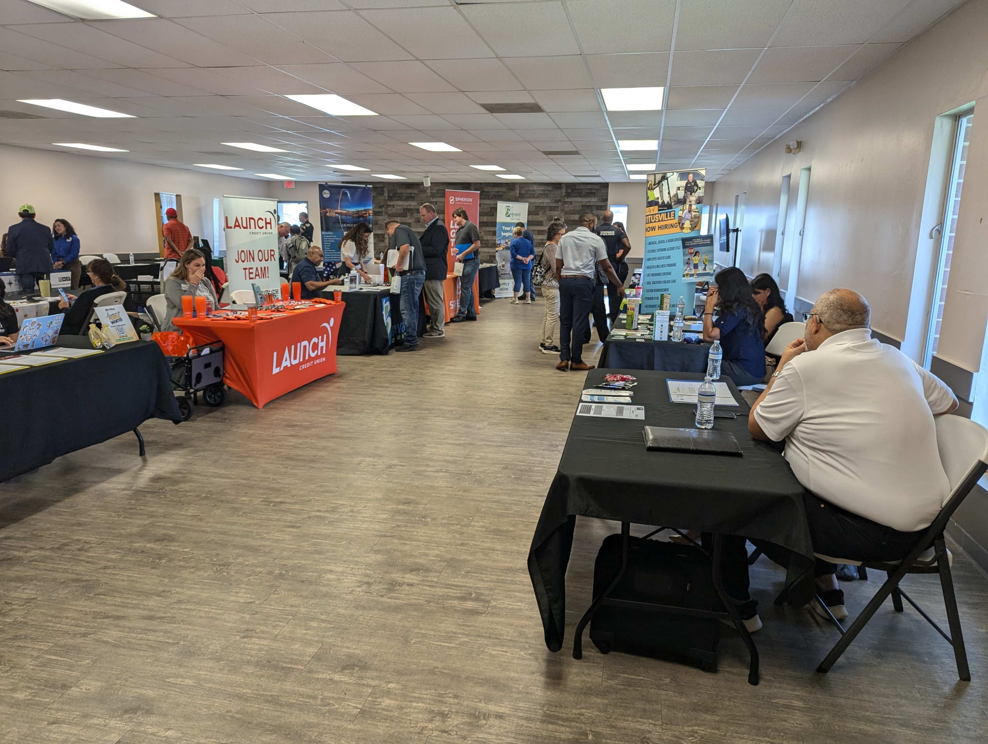 The image shows a room filled with people attending a job fair, featuring various booths displaying promotional materials and banners.