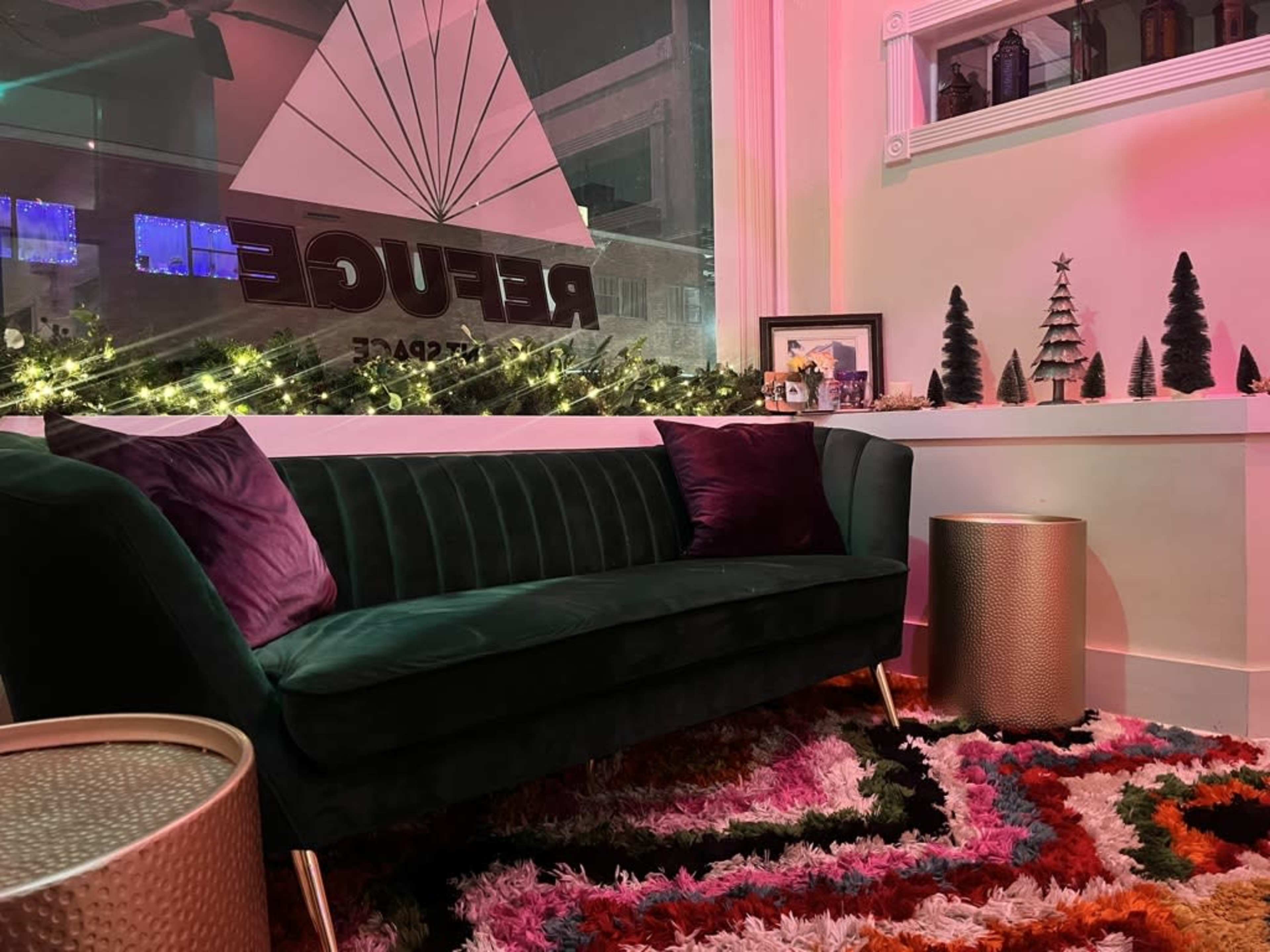 The image shows a cozy corner with a green velvet sofa, a colorful rug, and decorated shelves featuring small trees and twinkling lights.