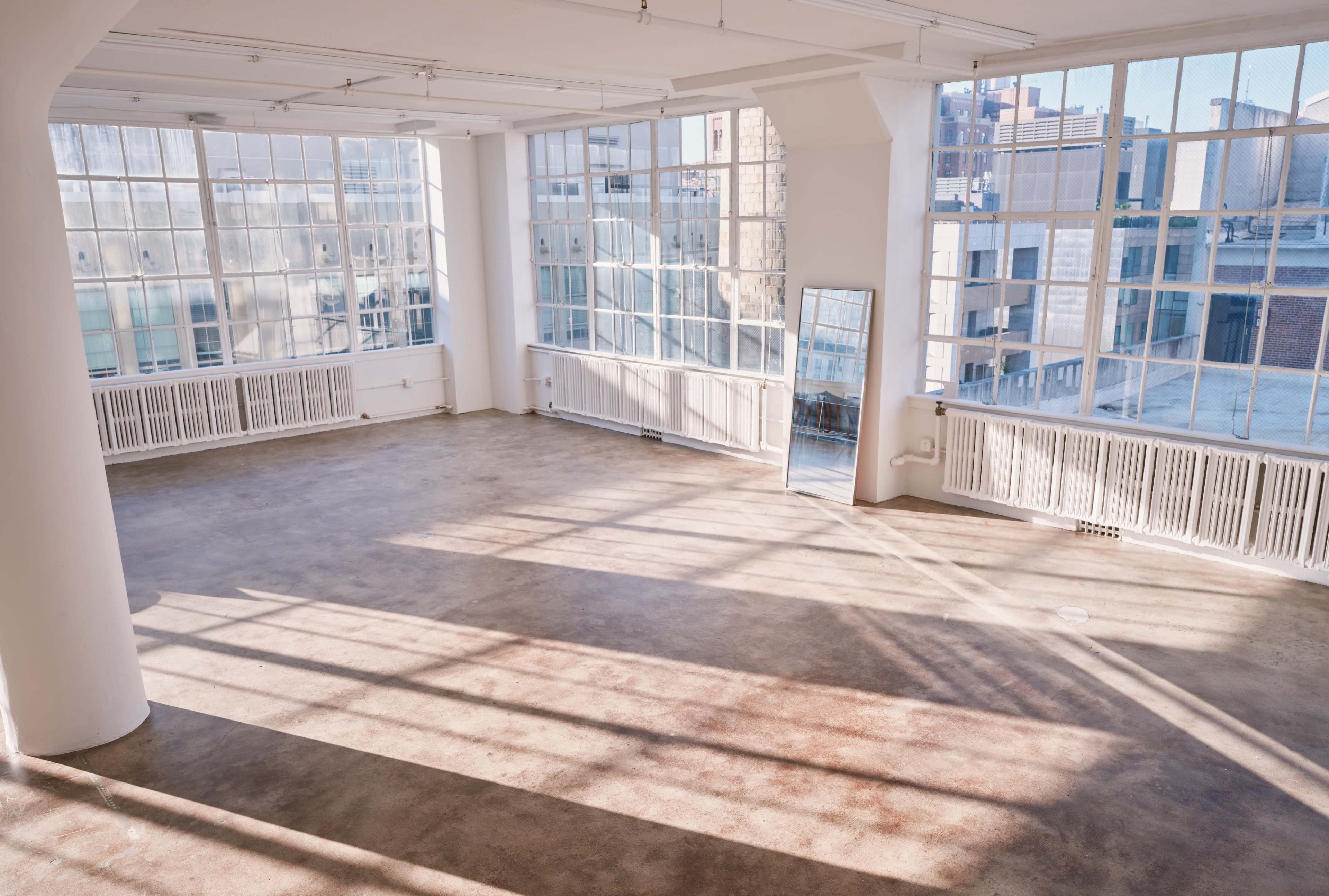 A spacious, empty room with large windows that allow natural light to flood in, casting shadows on the polished concrete floor.