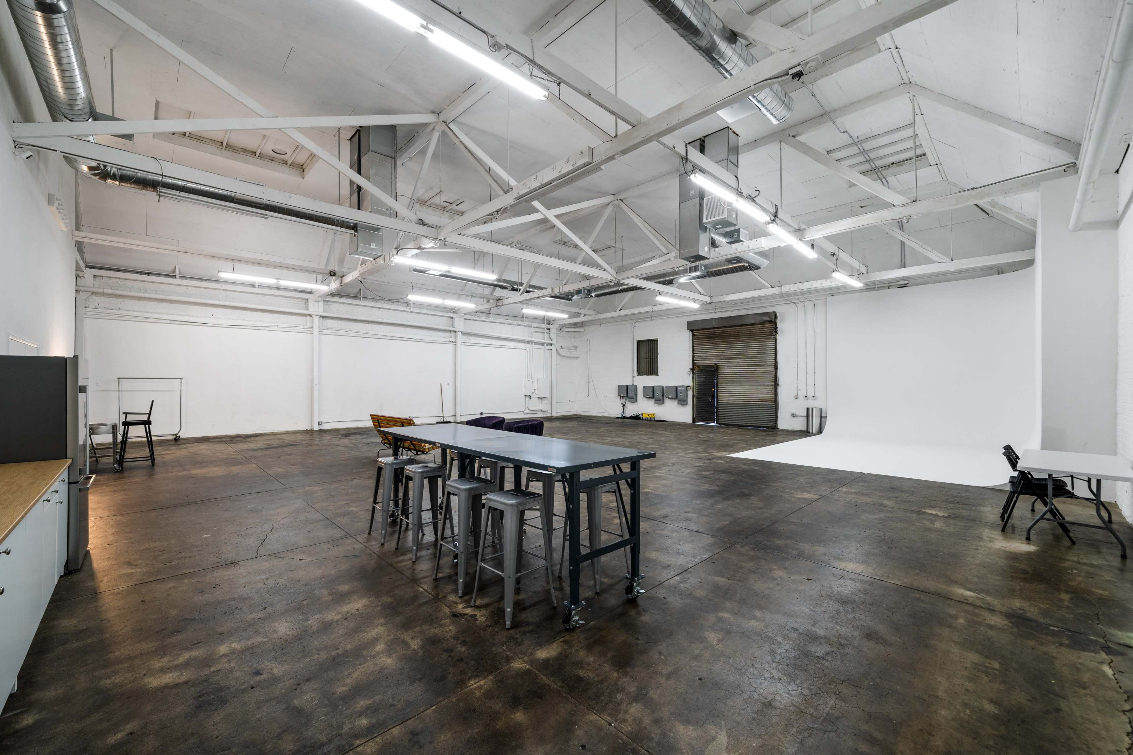 A spacious, industrial-style room with white walls, high ceilings, and a polished concrete floor, featuring a central table surrounded by stools and several empty chairs.