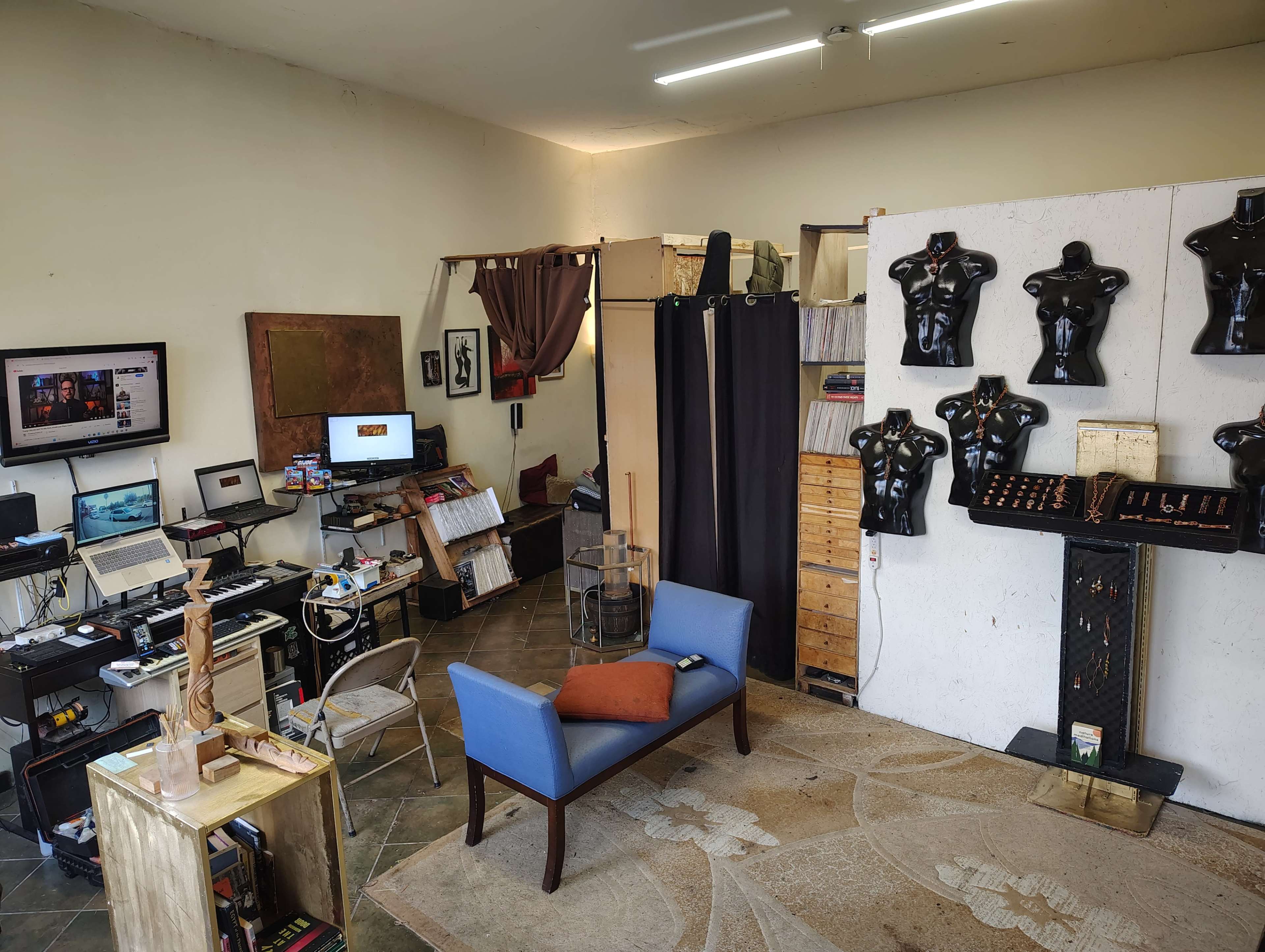 The image shows a spacious room containing a mix of office equipment, a seating area with a blue chair and an orange pillow, and several black dressmaker mannequins mounted on a wall.