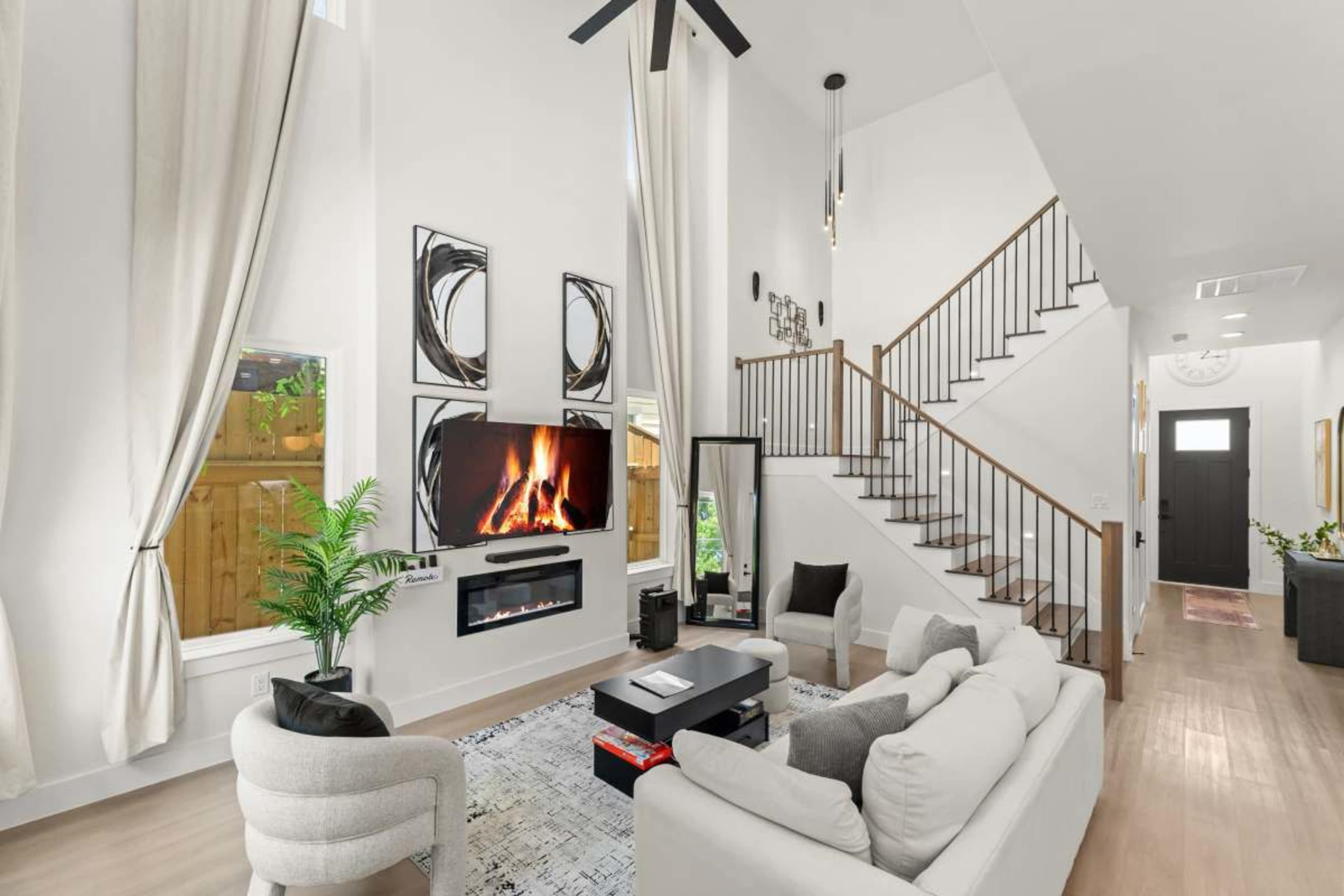 The image shows a modern living room with a large window, a television mounted on the wall above an electric fireplace, and a staircase leading to the upper level.