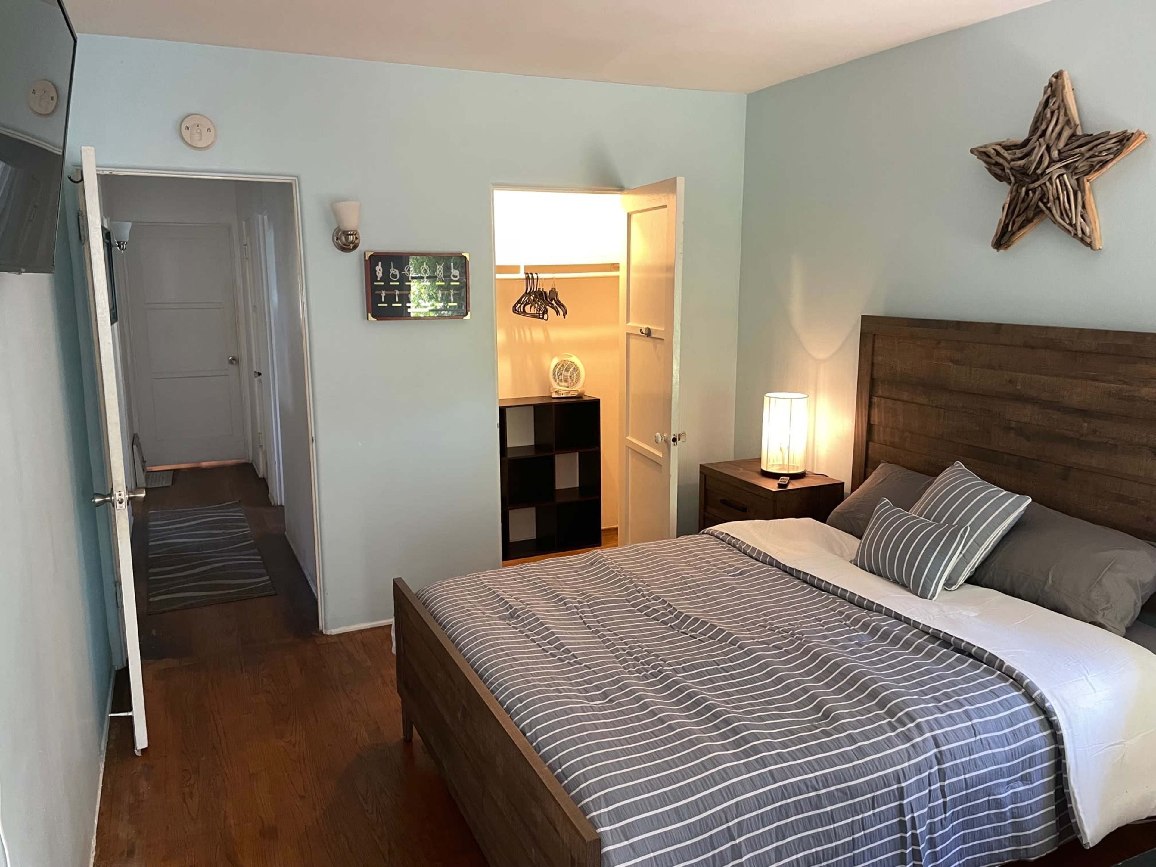 The image shows a small bedroom featuring a bed with grey bedding, a bedside lamp, and a closet with an open door, leading to a hallway.