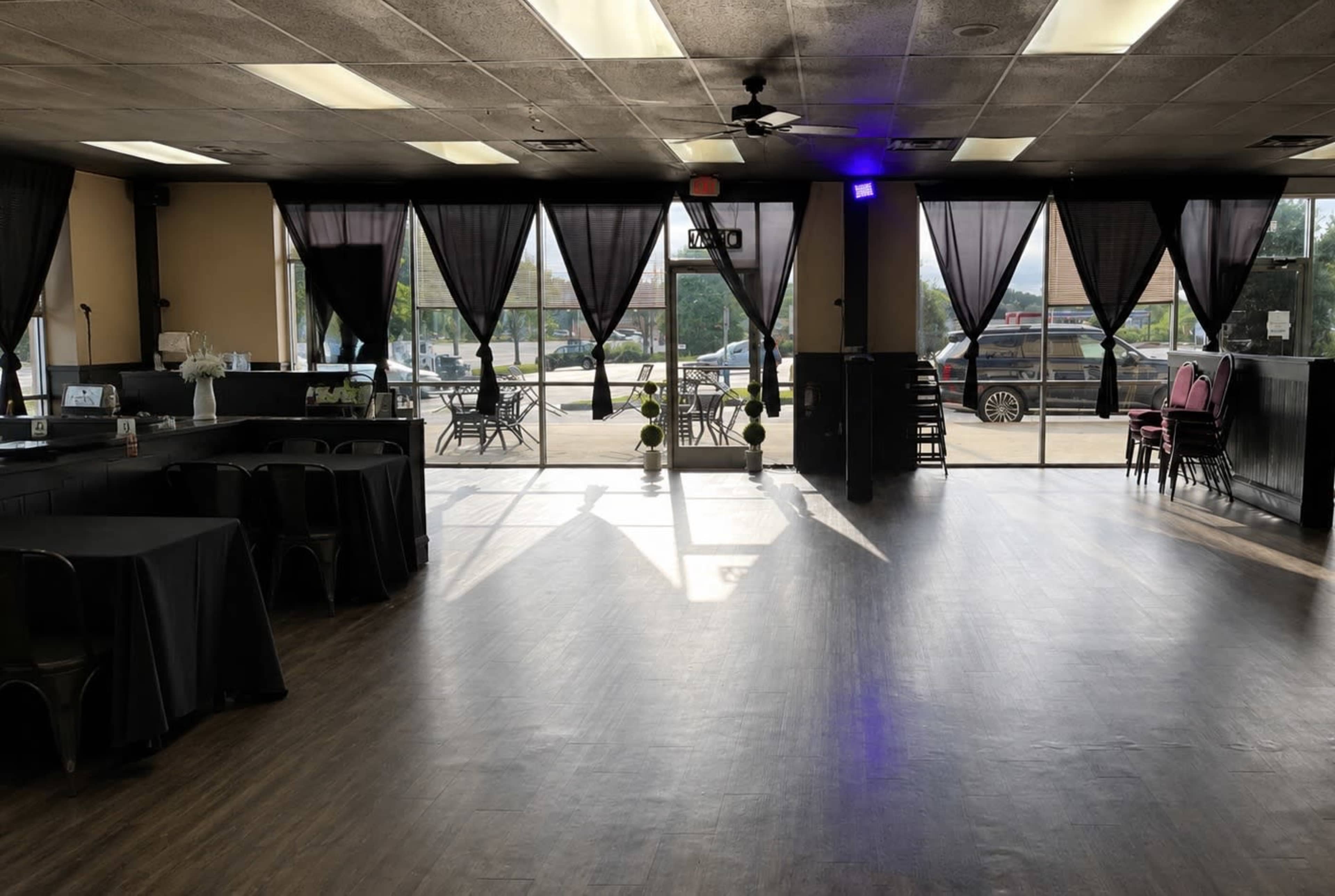 The image shows an interior space with large windows, dark tablecloth-covered tables, and black chairs, illuminated by natural light.