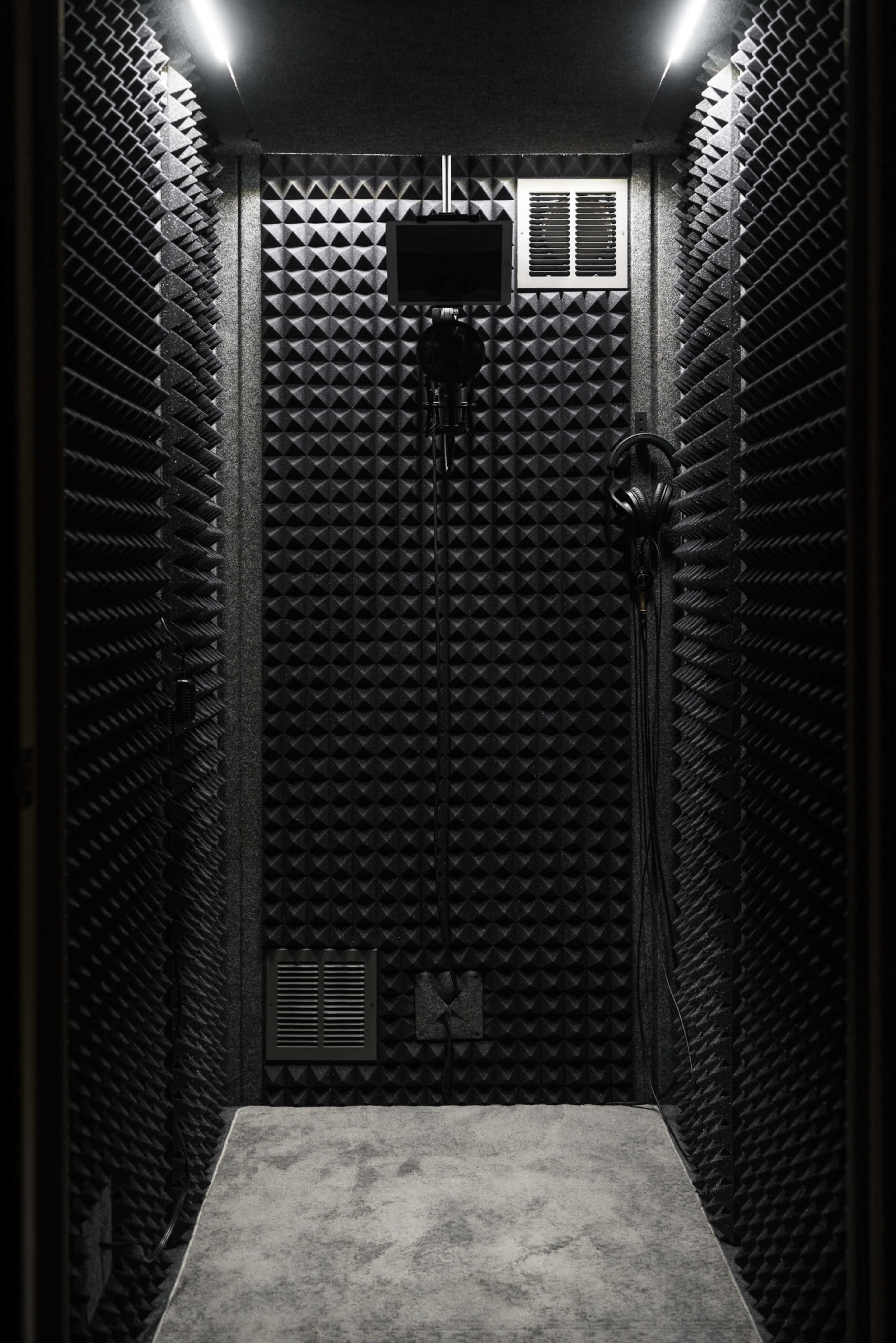 The image shows a small soundproof booth with textured acoustic panels on the walls and a microphone setup inside.
