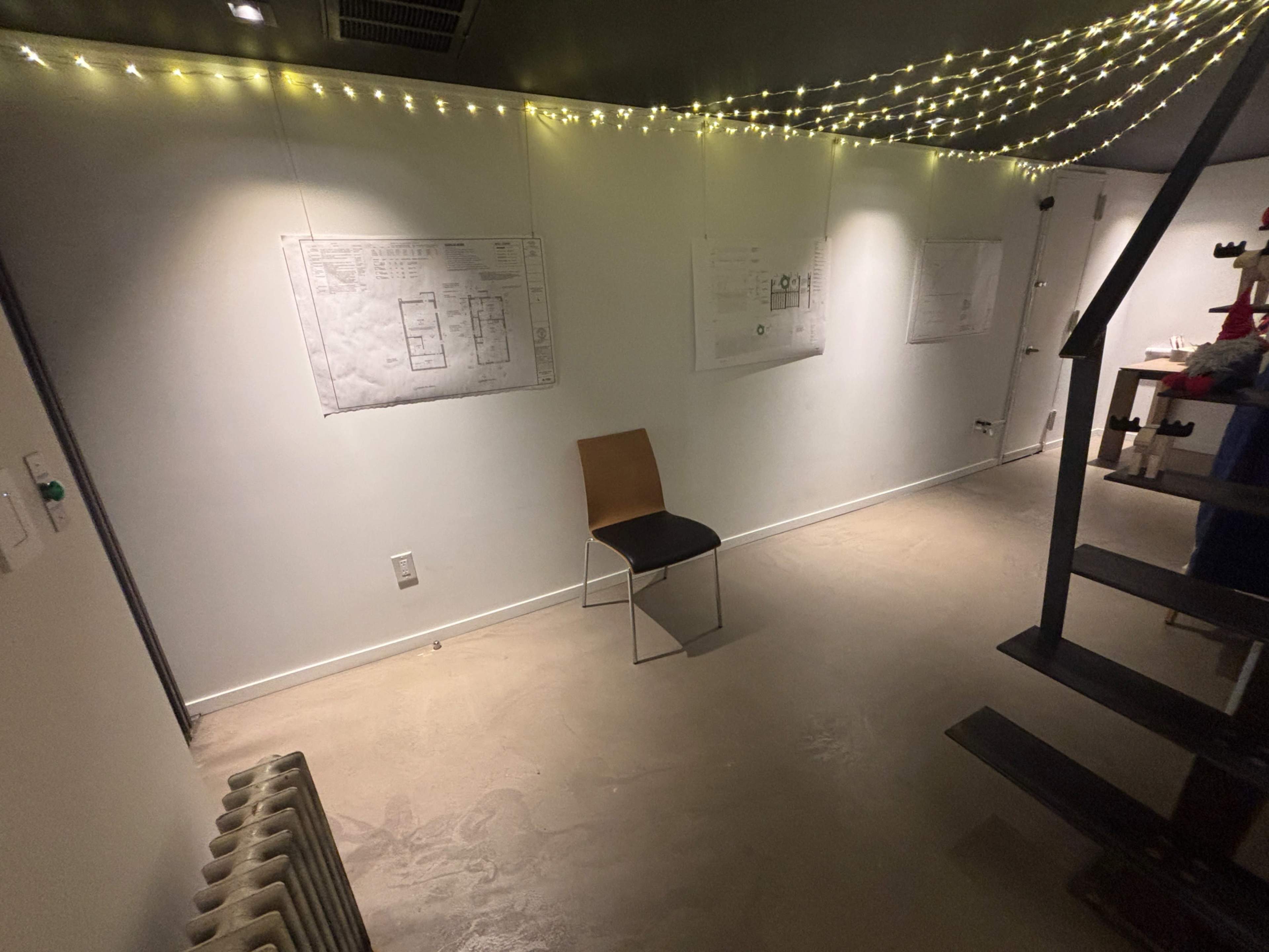 The image shows an empty room with a single chair, illuminated by string lights, and architectural plans hanging on the walls.