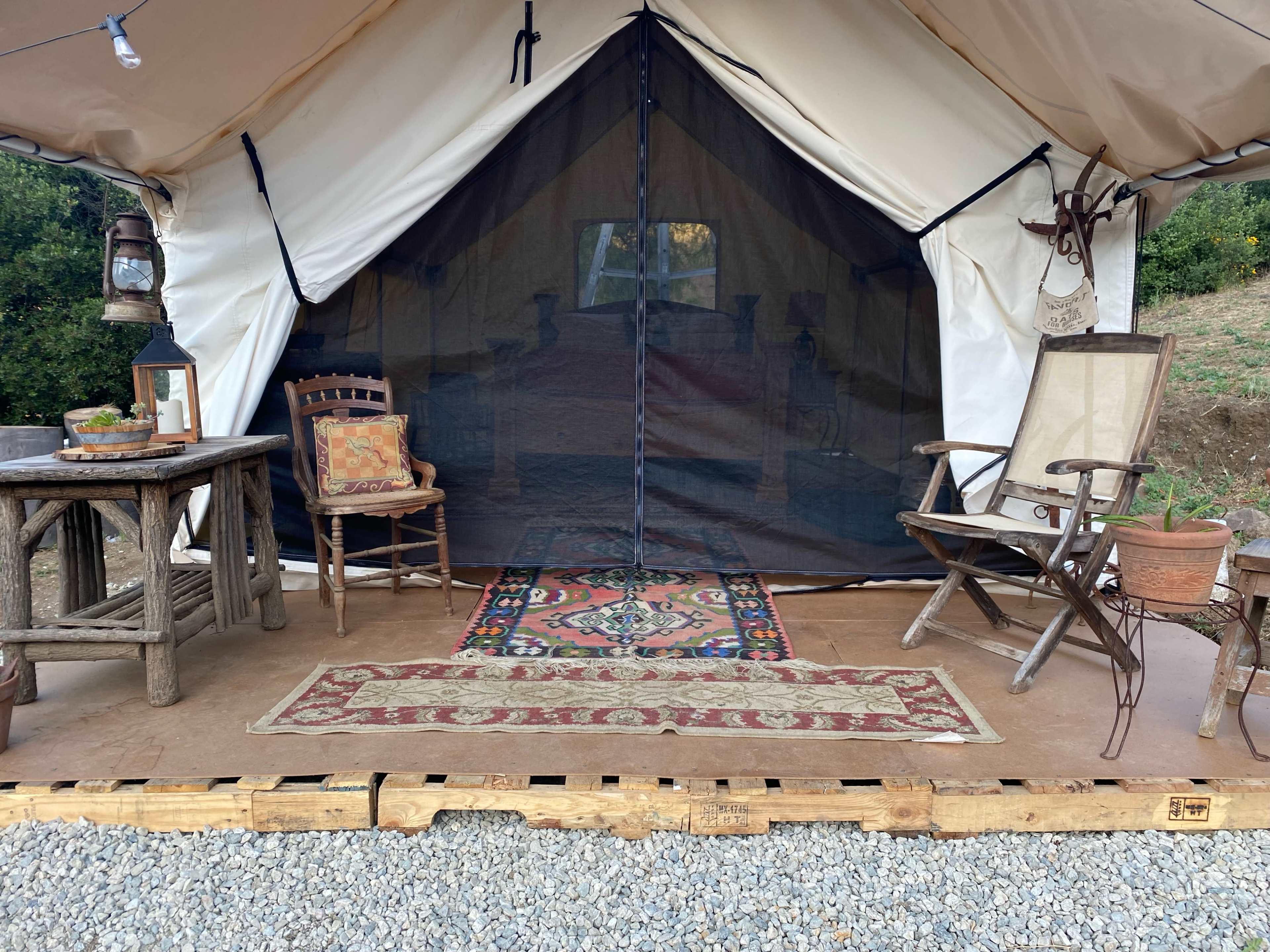 A tent entrance with wooden furniture, a rug, and a lantern arranged on a gravel surface.