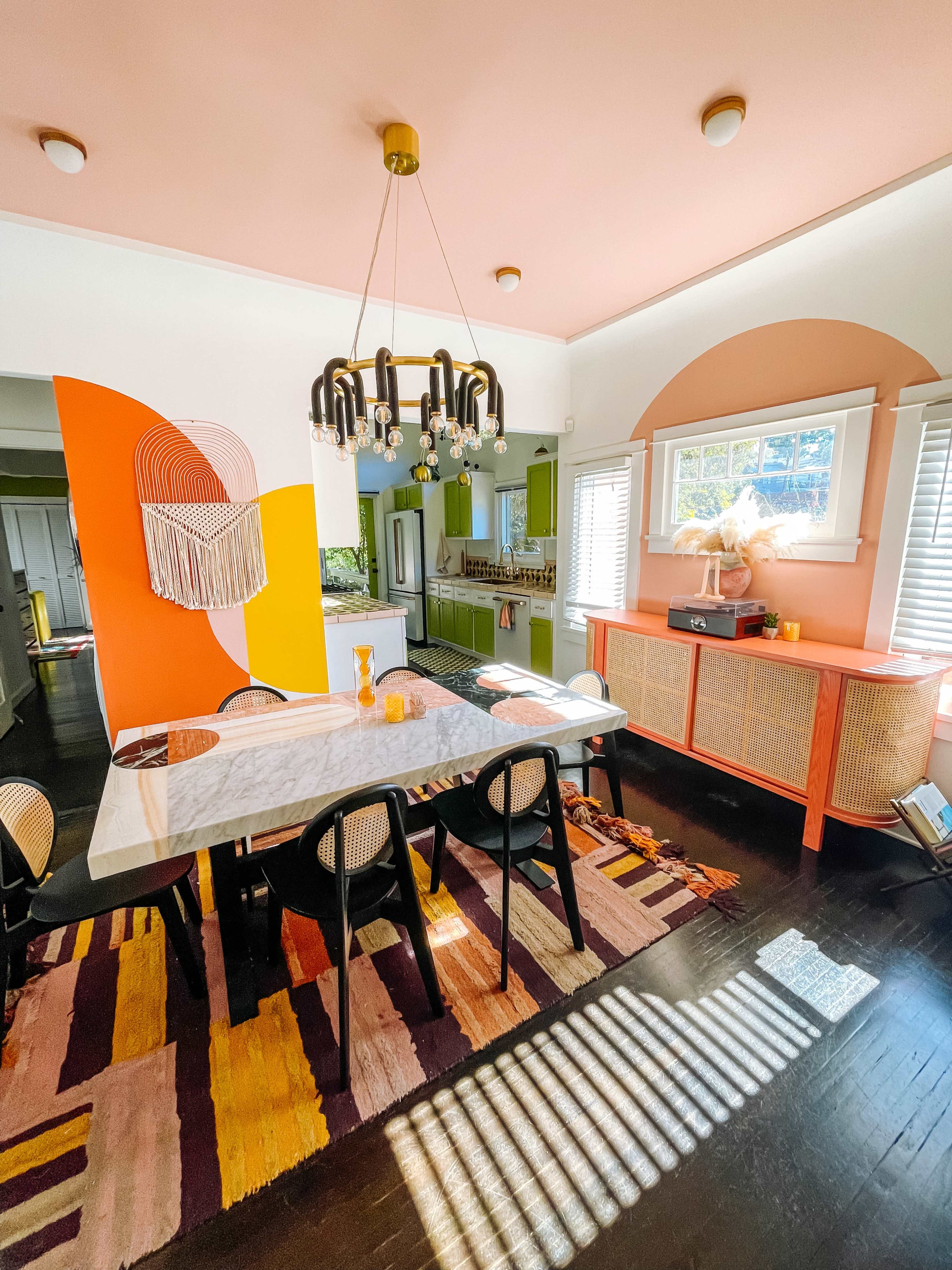 A dining area featuring a marble table with black chairs, a colorful accent wall, and warm natural light streaming through the windows.