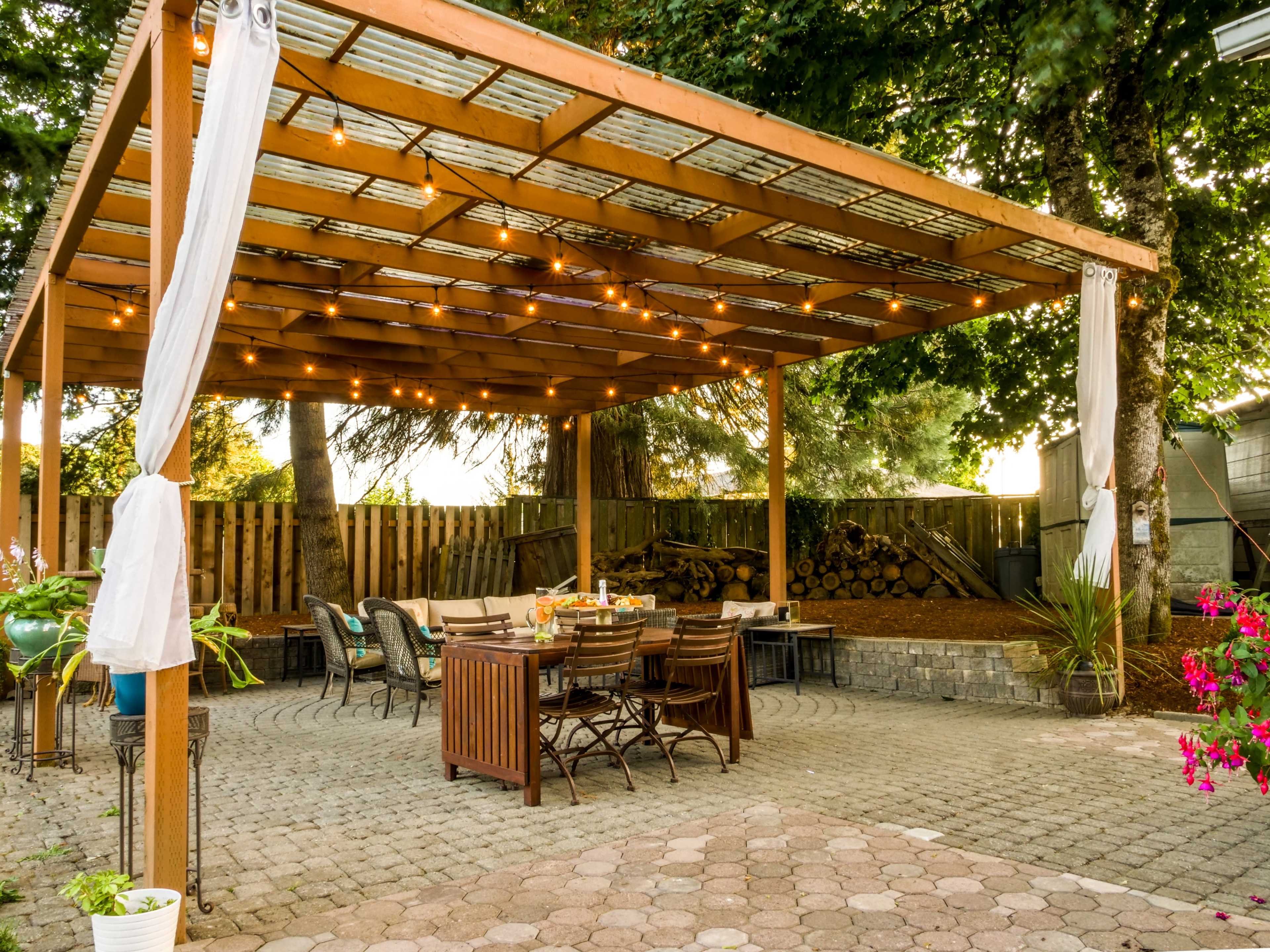 A wooden pergola with string lights over a brick patio featuring a dining table and chairs, surrounded by trees and a wooden fence.