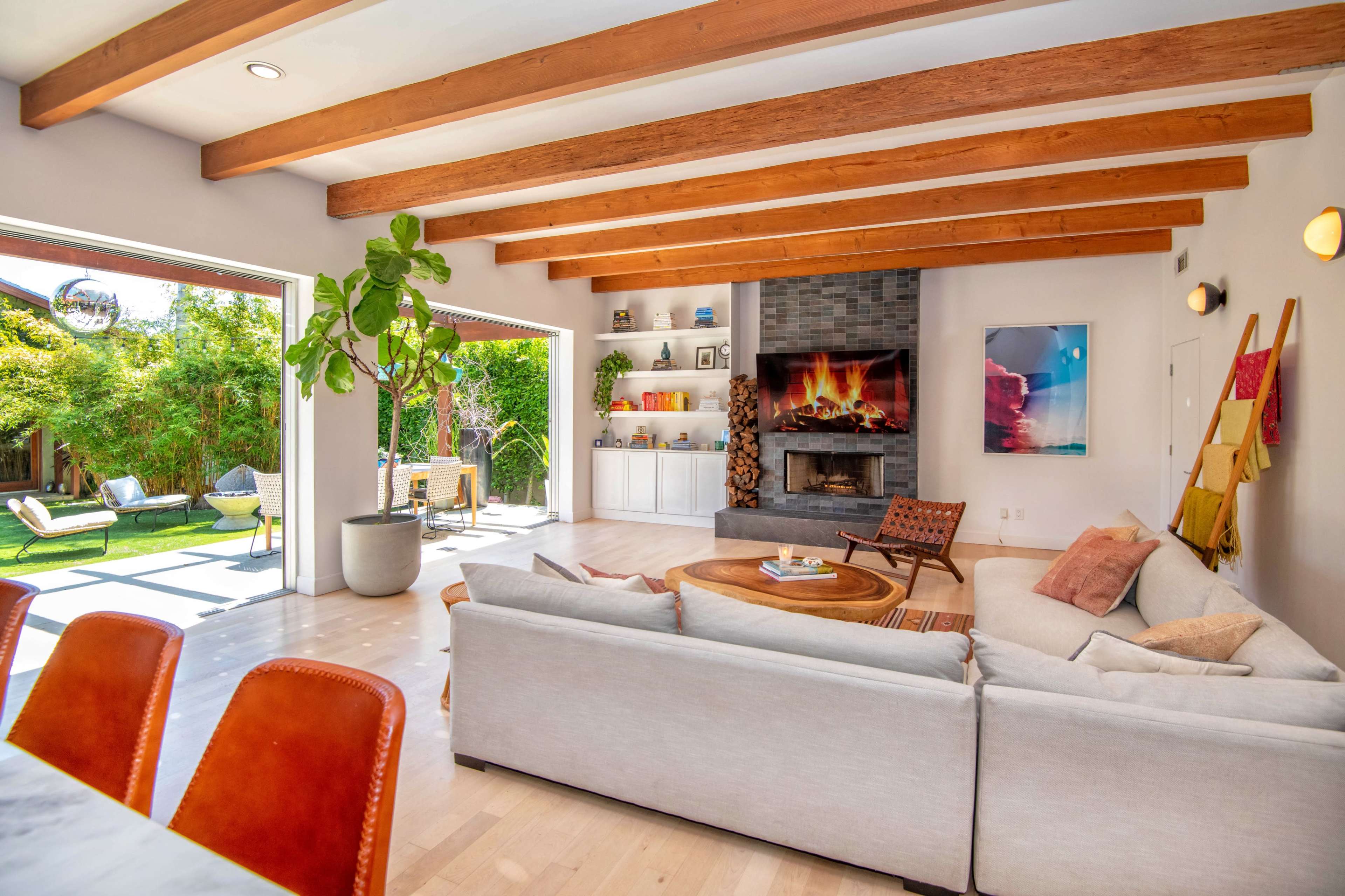 A modern living room features a light-colored sofa, a circular wooden coffee table, and a stone fireplace, with large openings leading to an outdoor patio and garden.