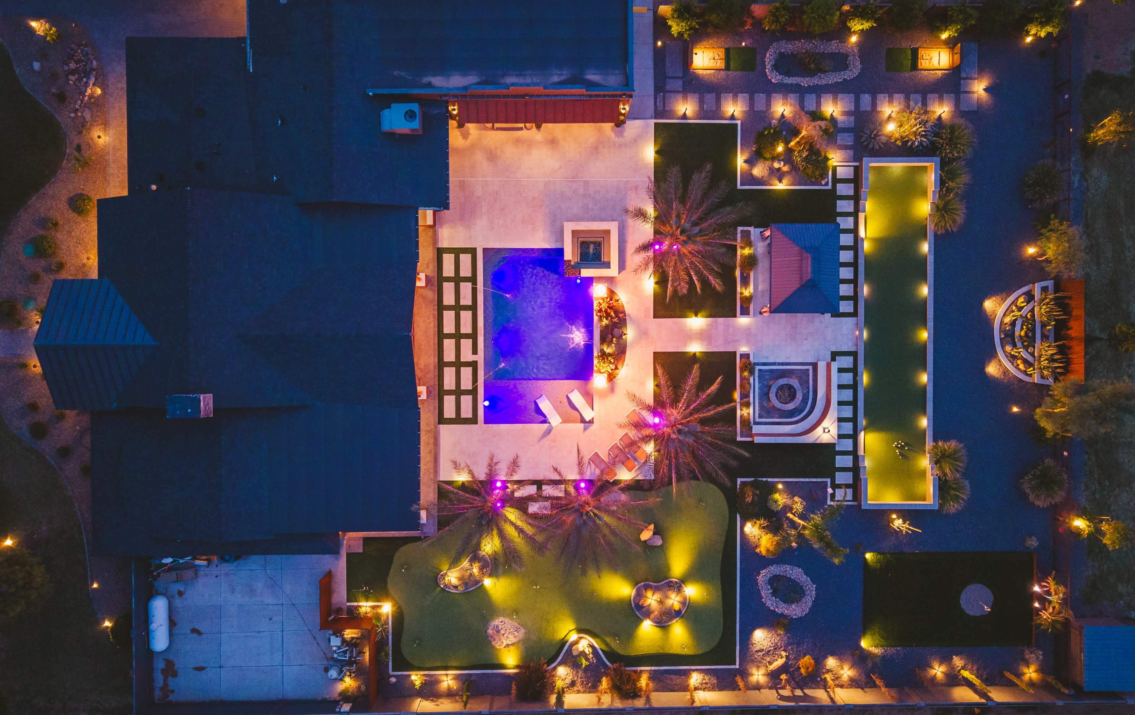 An aerial view of a landscaped backyard featuring a swimming pool, hot tub, and various illuminated outdoor spaces surrounded by palm trees and decorative lighting.