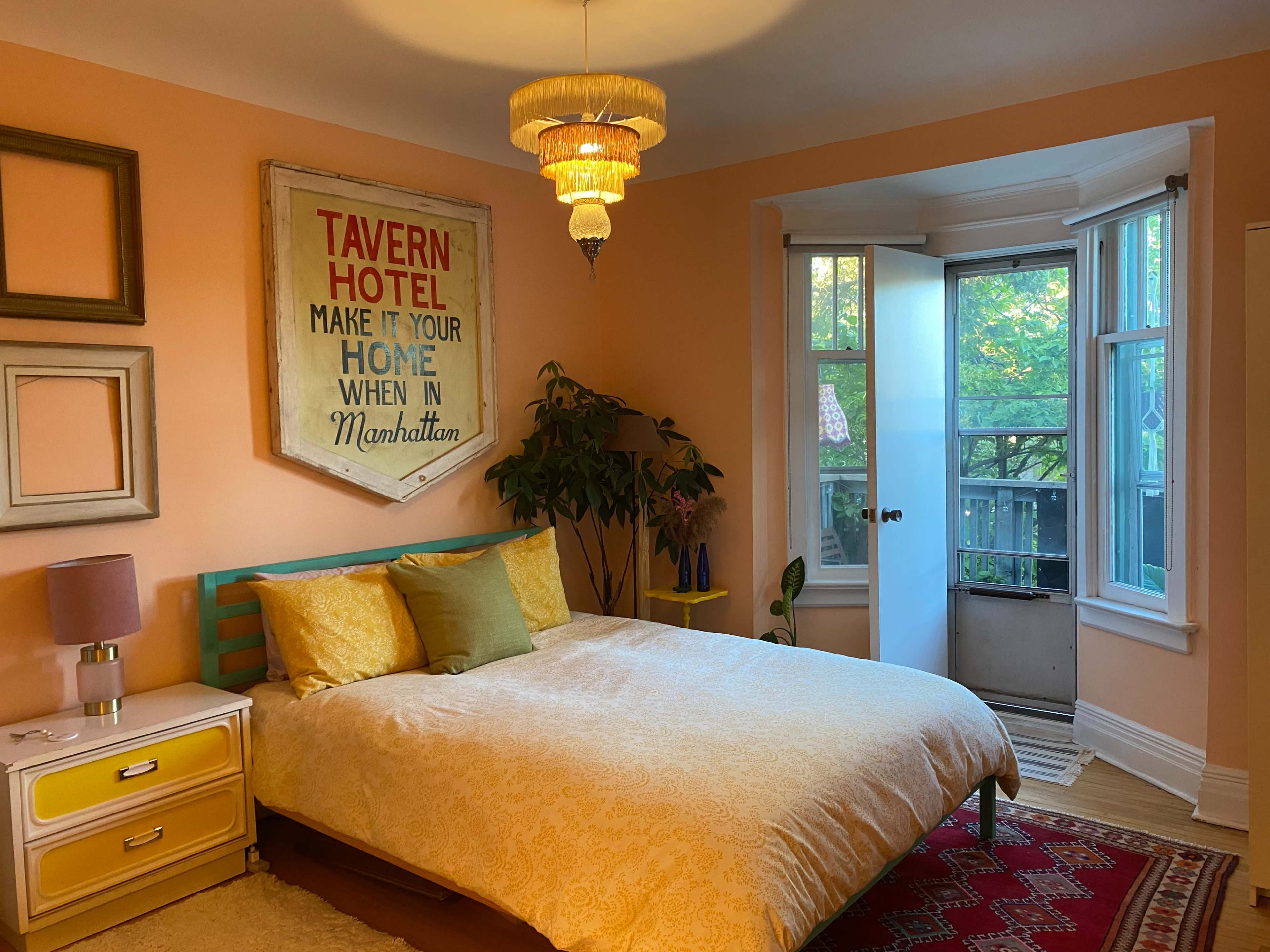 A bedroom features a bed with a patterned comforter, a vintage sign above it, pastel-colored walls, and a window that opens to a view of greenery.