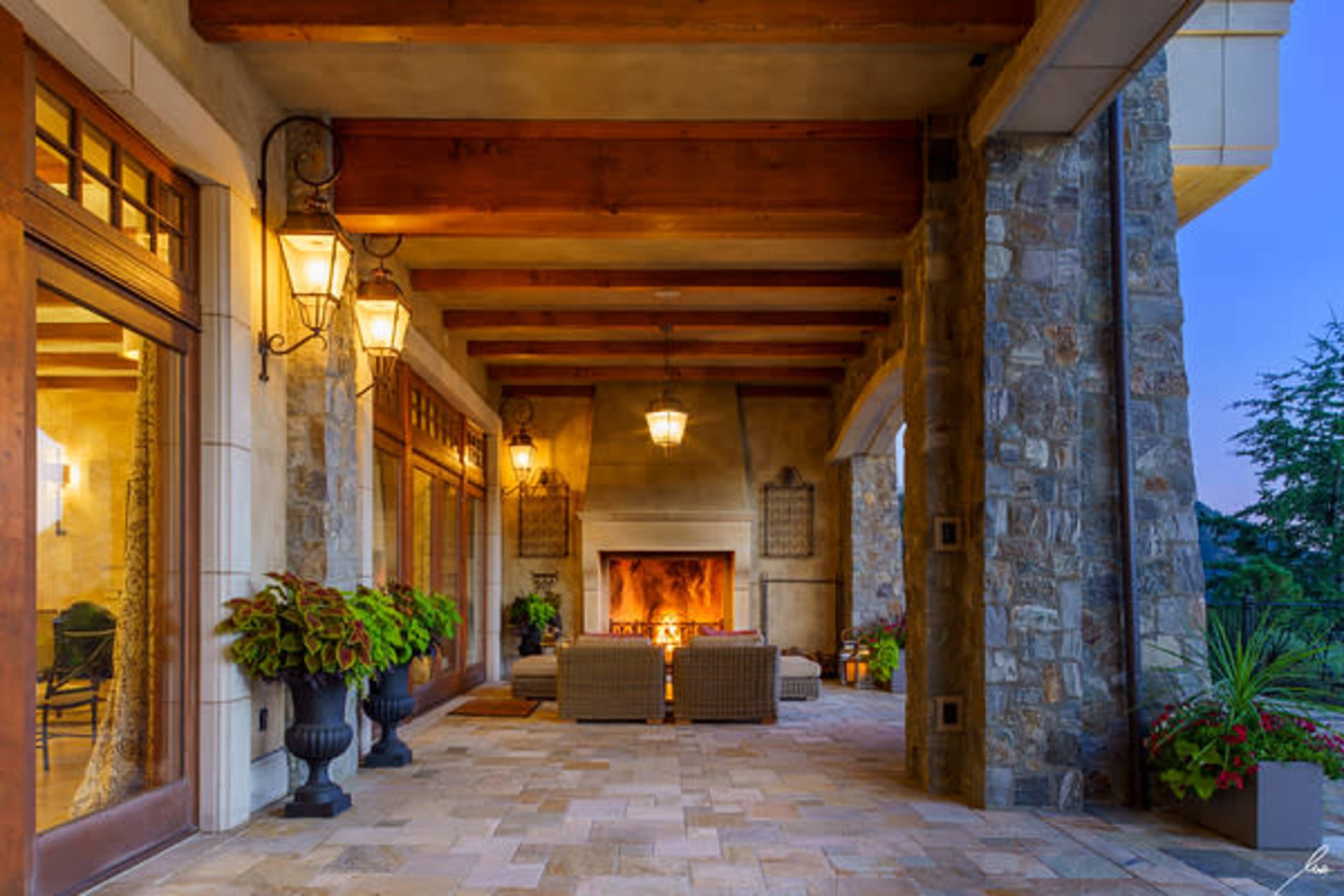 A covered patio with stone walls, wooden beams, and a cozy seating area arranged around a fireplace, surrounded by potted plants.
