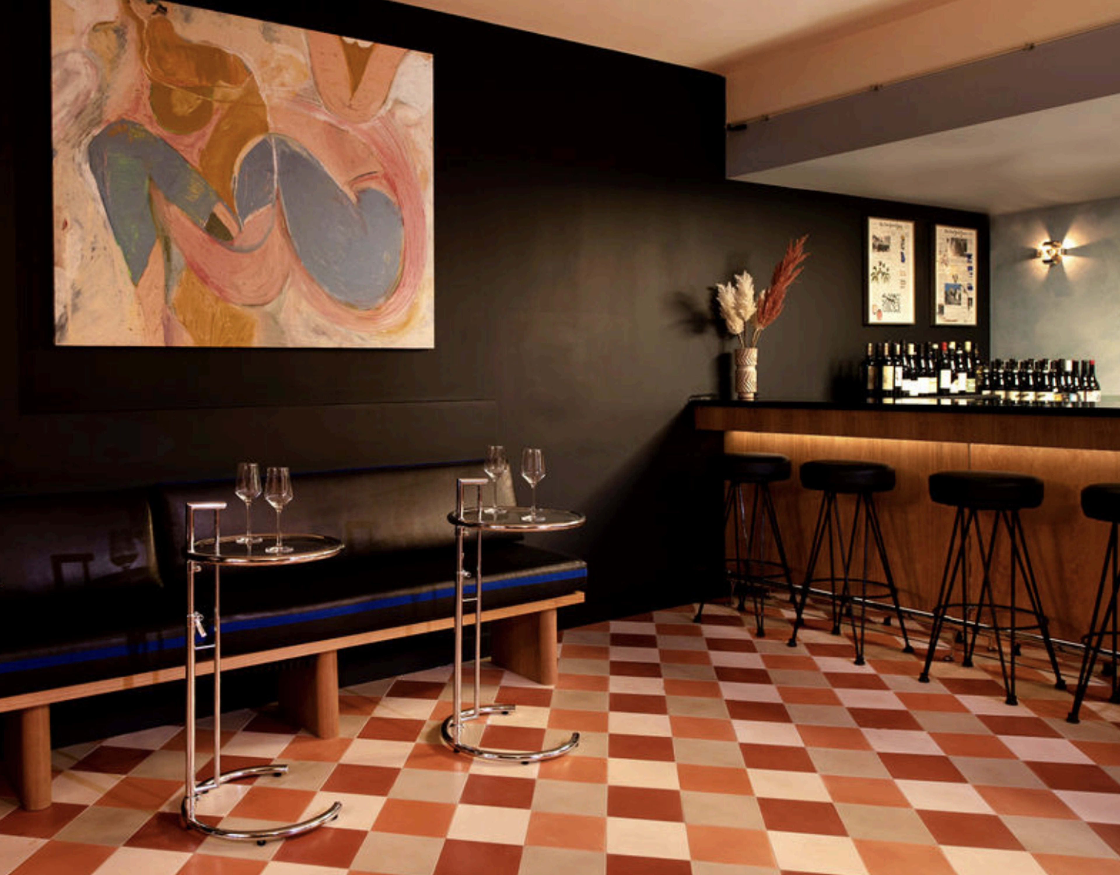 The image shows a stylish bar area with a dark wall, colorful abstract art, and two modern glass-top tables in front of a wooden bar lined with wine bottles.