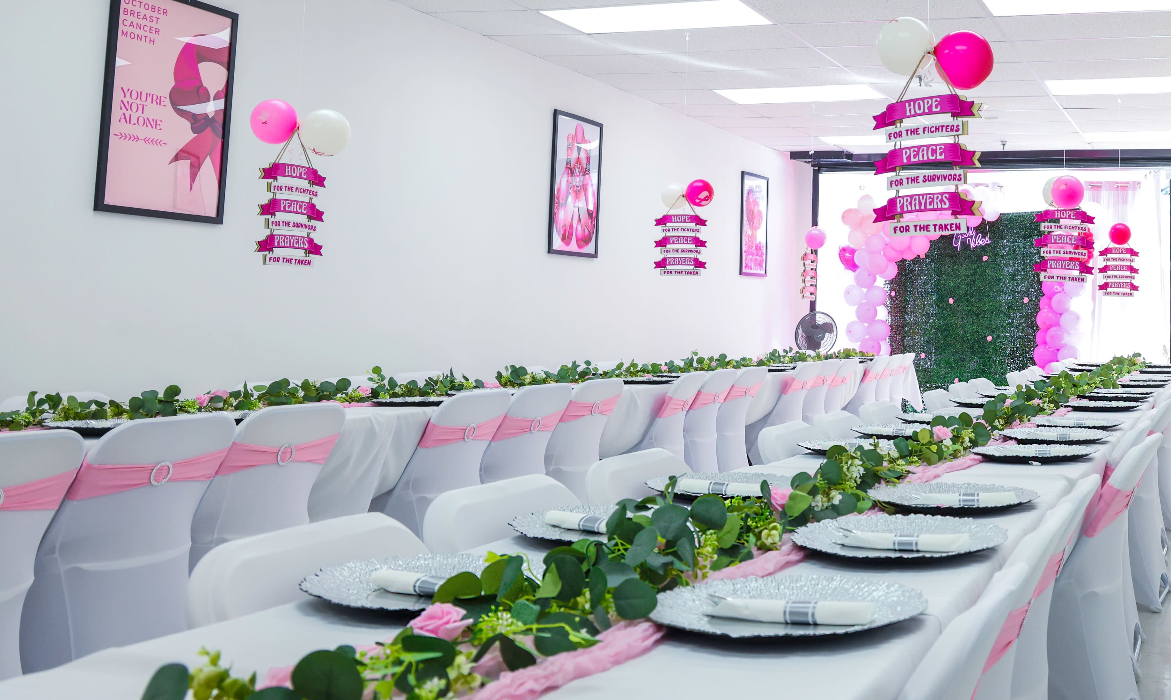 A long banquet table is decorated with greenery and pink accents, set in a bright room adorned with motivational posters and pink balloons.