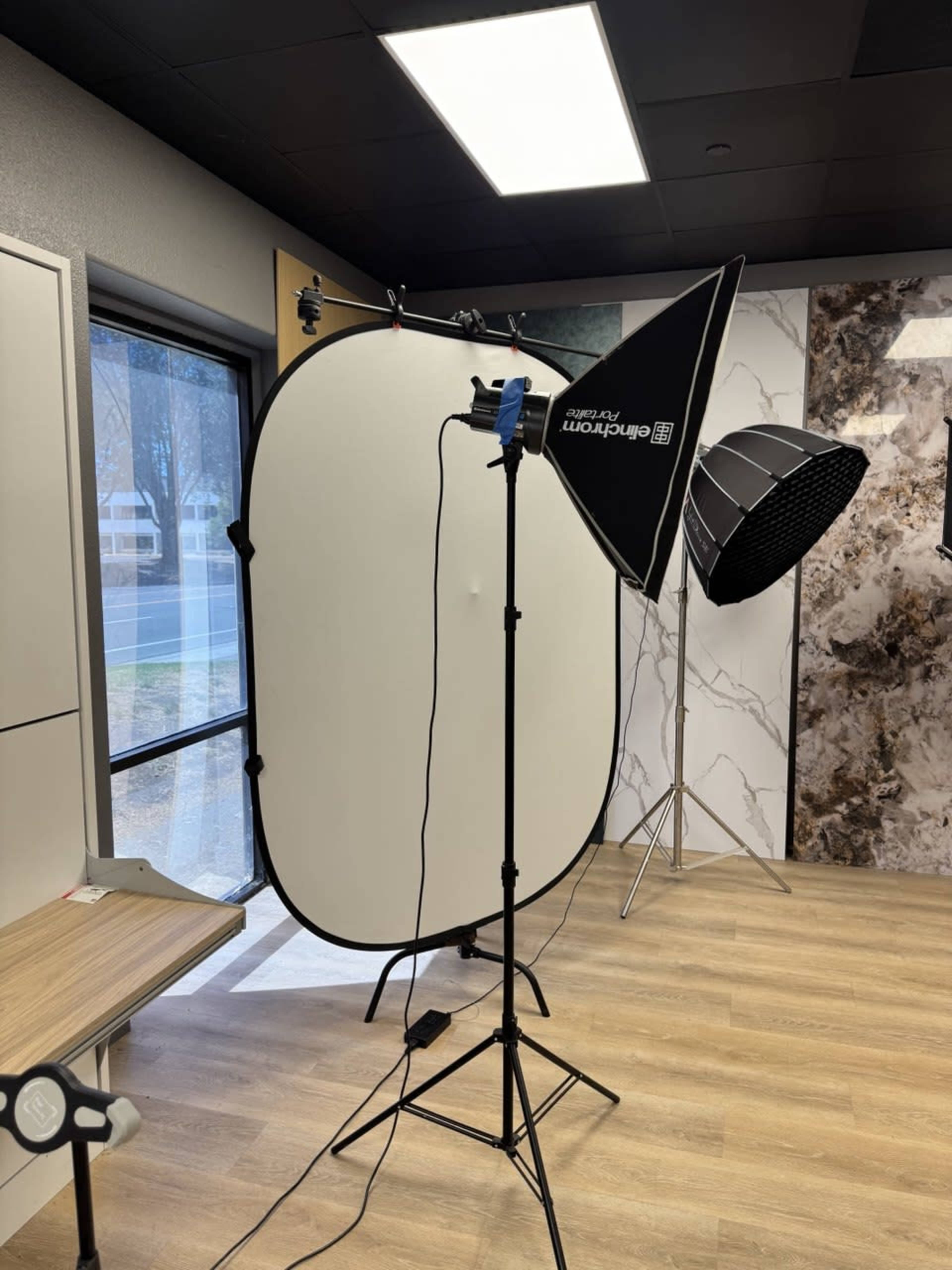 The image shows a photography setup with a large light reflector, two softboxes, and a window in a well-lit room featuring wood flooring.
