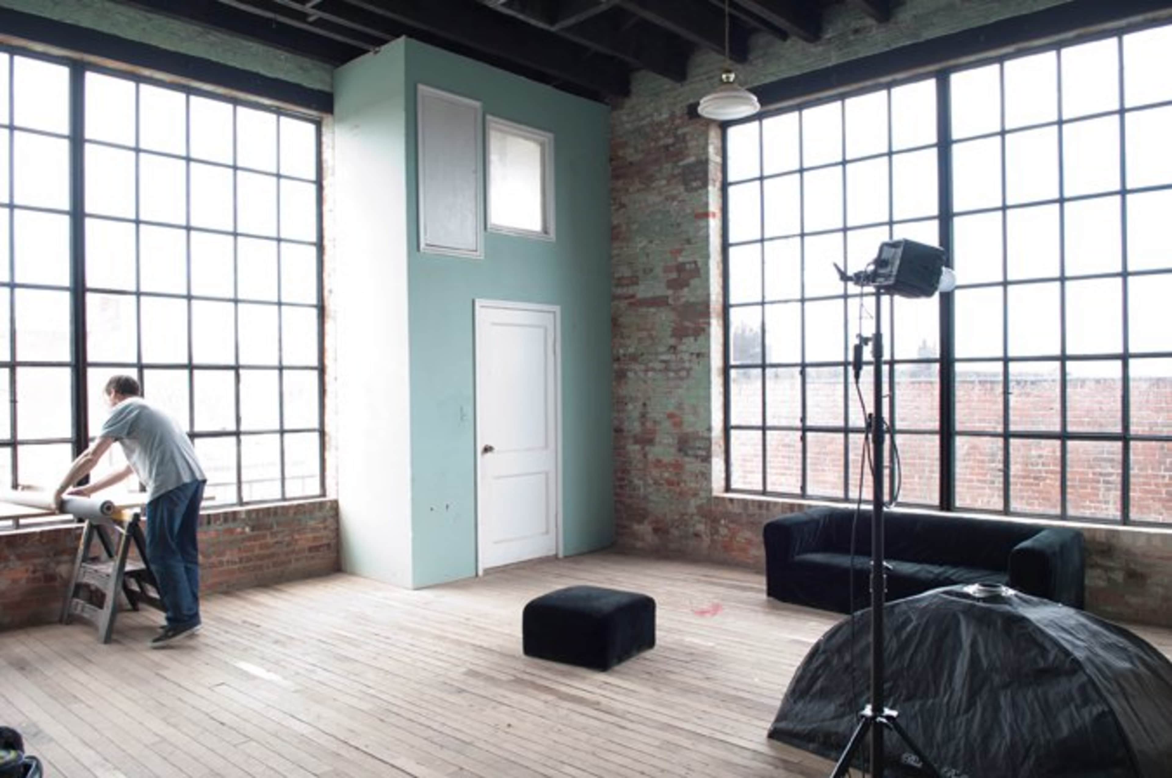 A person is working at a table in a spacious, industrial-style room with large windows and exposed brick walls.