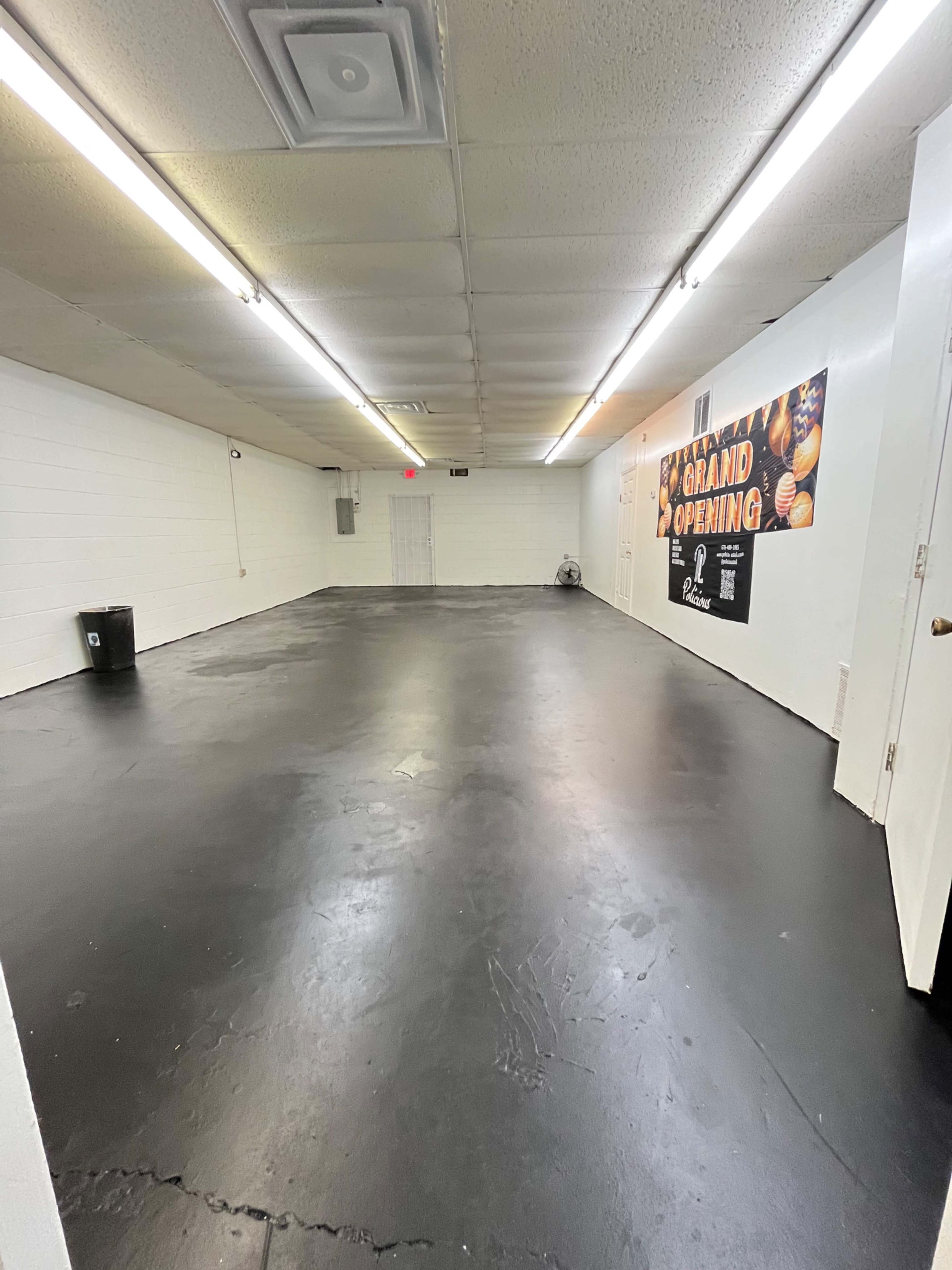 An empty room with a black floor, white walls, and overhead fluorescent lighting, featuring a promotional poster for a grand opening on one wall.