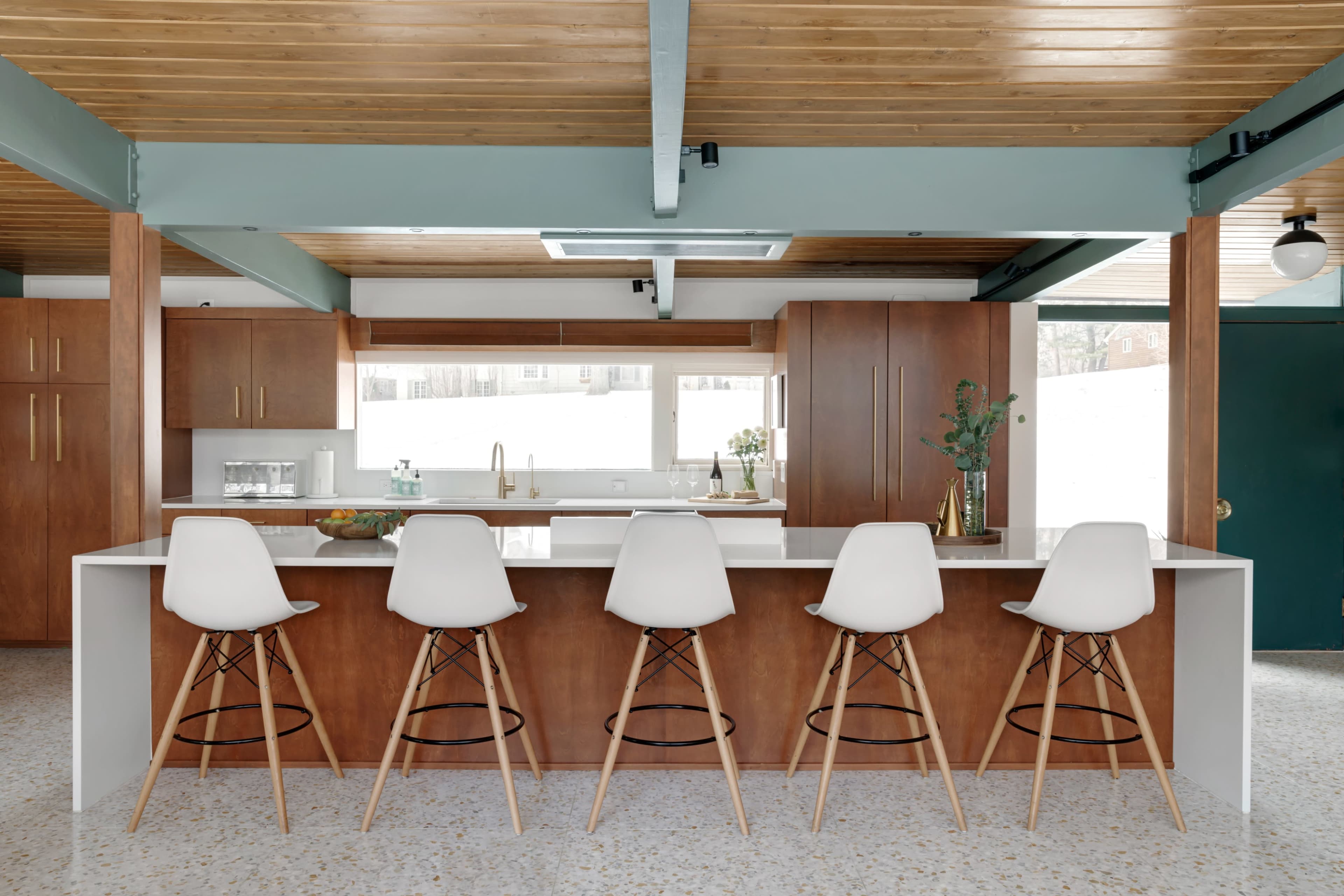 A modern kitchen features a white island with five wooden bar stools and wood-paneled cabinetry.