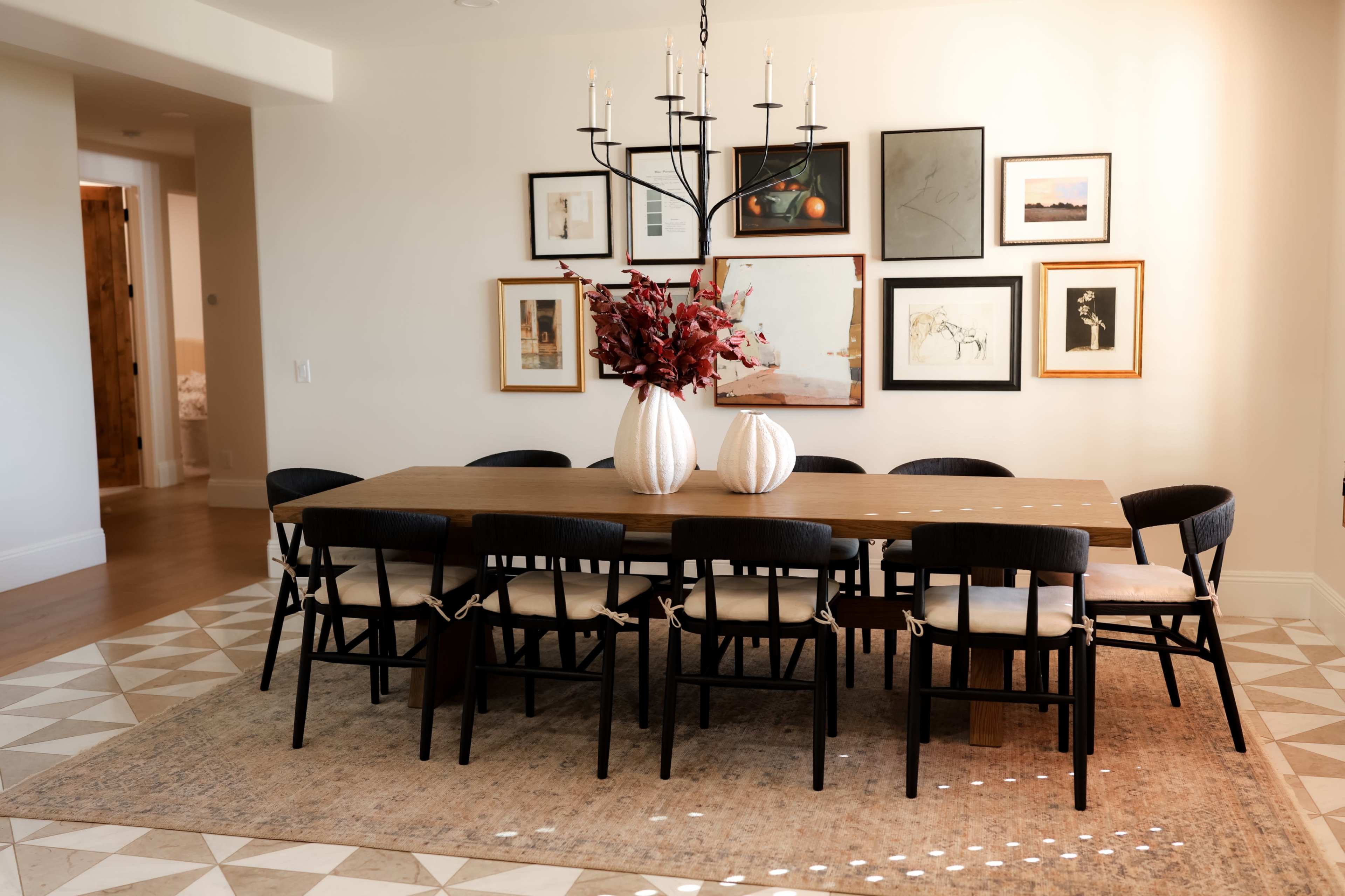 A long wooden dining table with black chairs is set under a chandelier, surrounded by framed artworks on the wall.