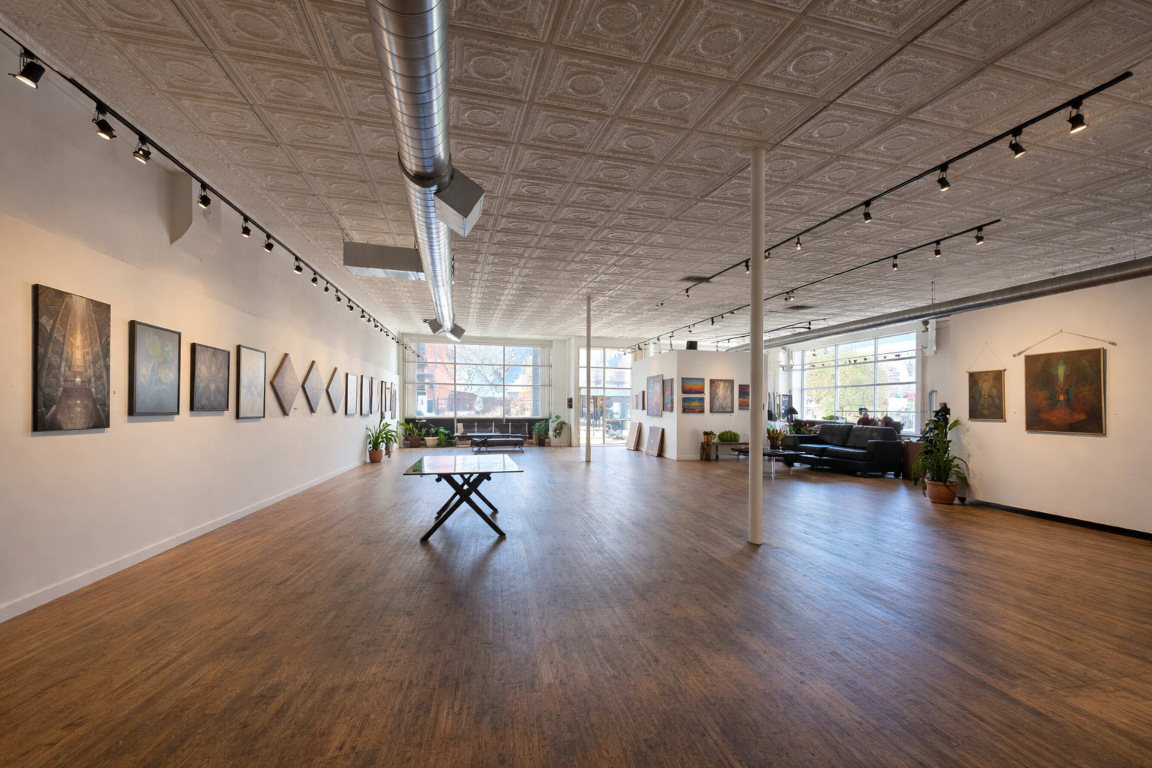 The image shows a spacious art gallery with various artworks displayed on the walls and a table in the center, surrounded by plants and natural light from large windows.