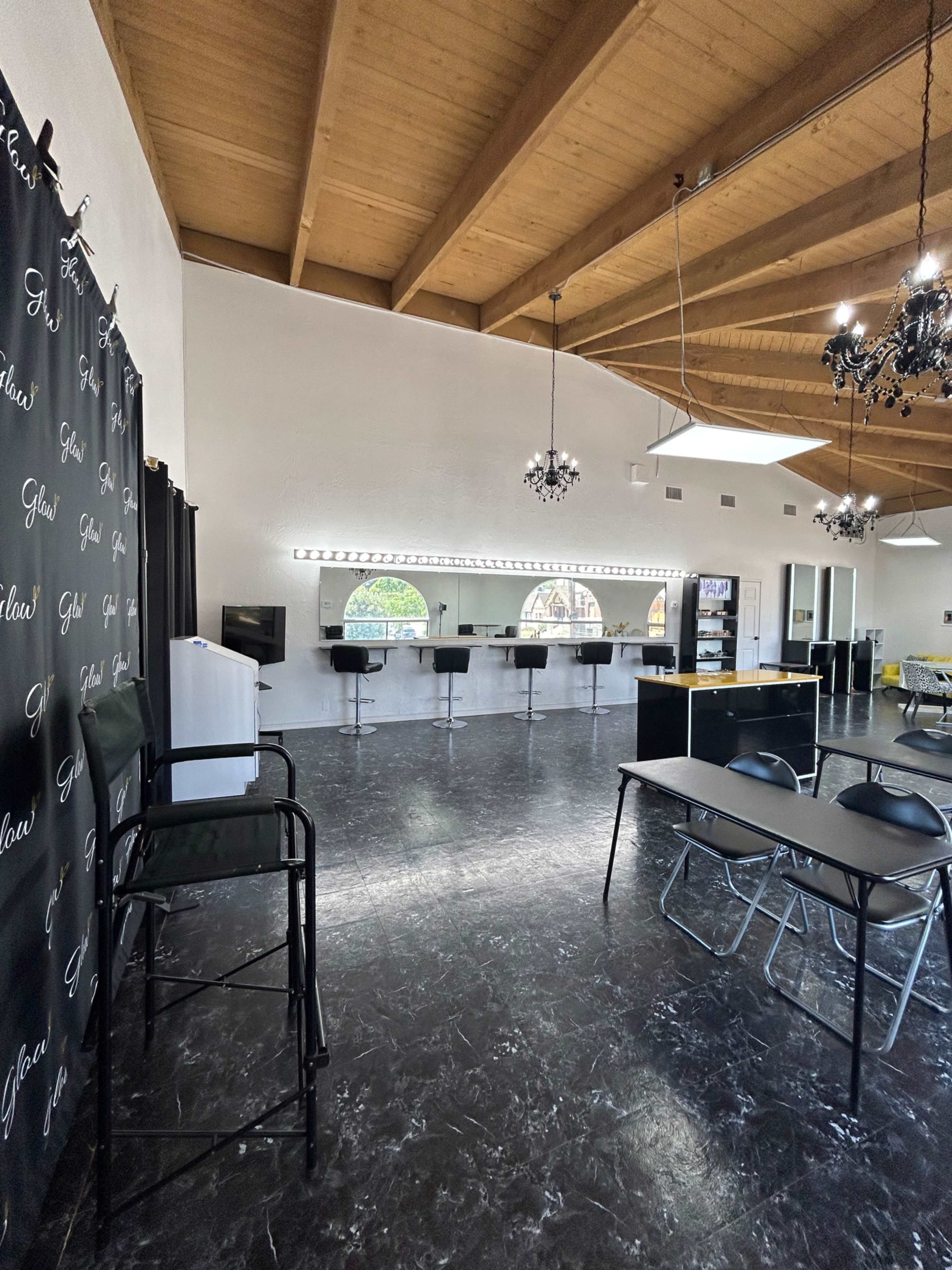 A spacious room with a high wooden ceiling, featuring several tables and chairs, a mirrored wall with lights, and a service counter against one side.