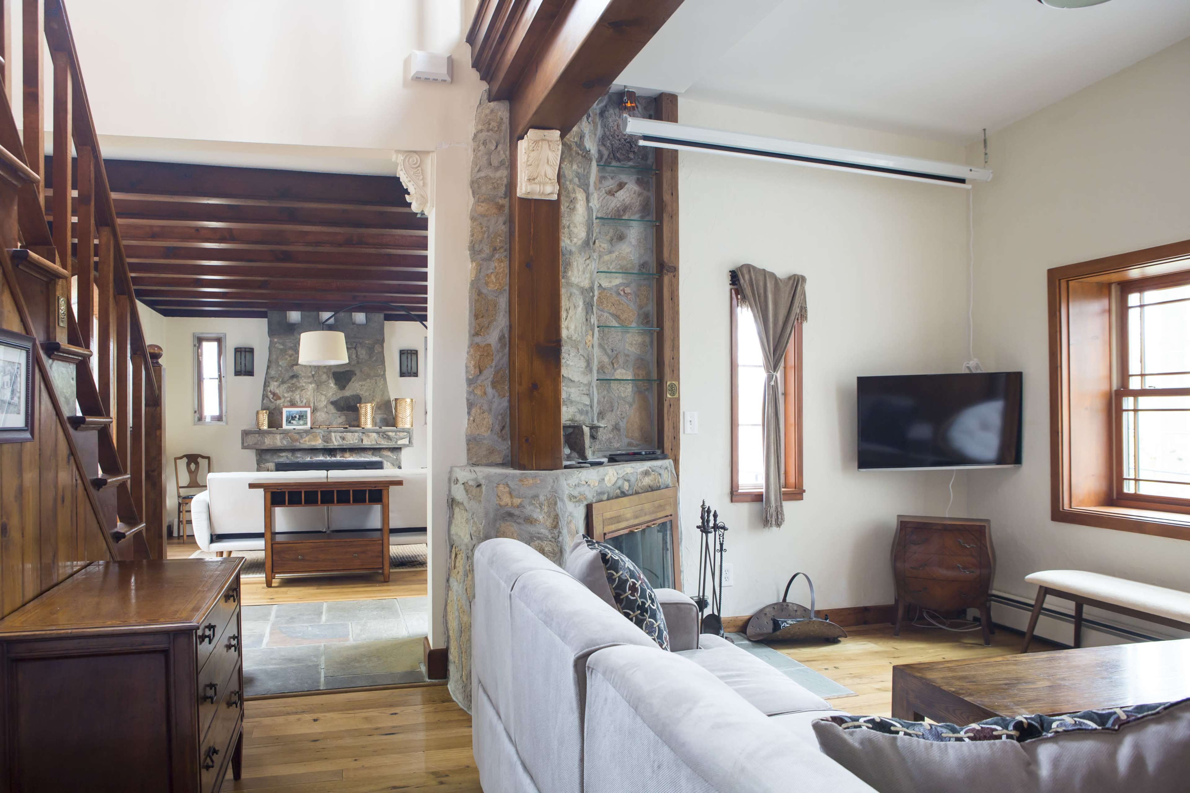 A cozy living space featuring wooden beams, a stone fireplace, and a connected kitchen area with a television mounted on the wall.
