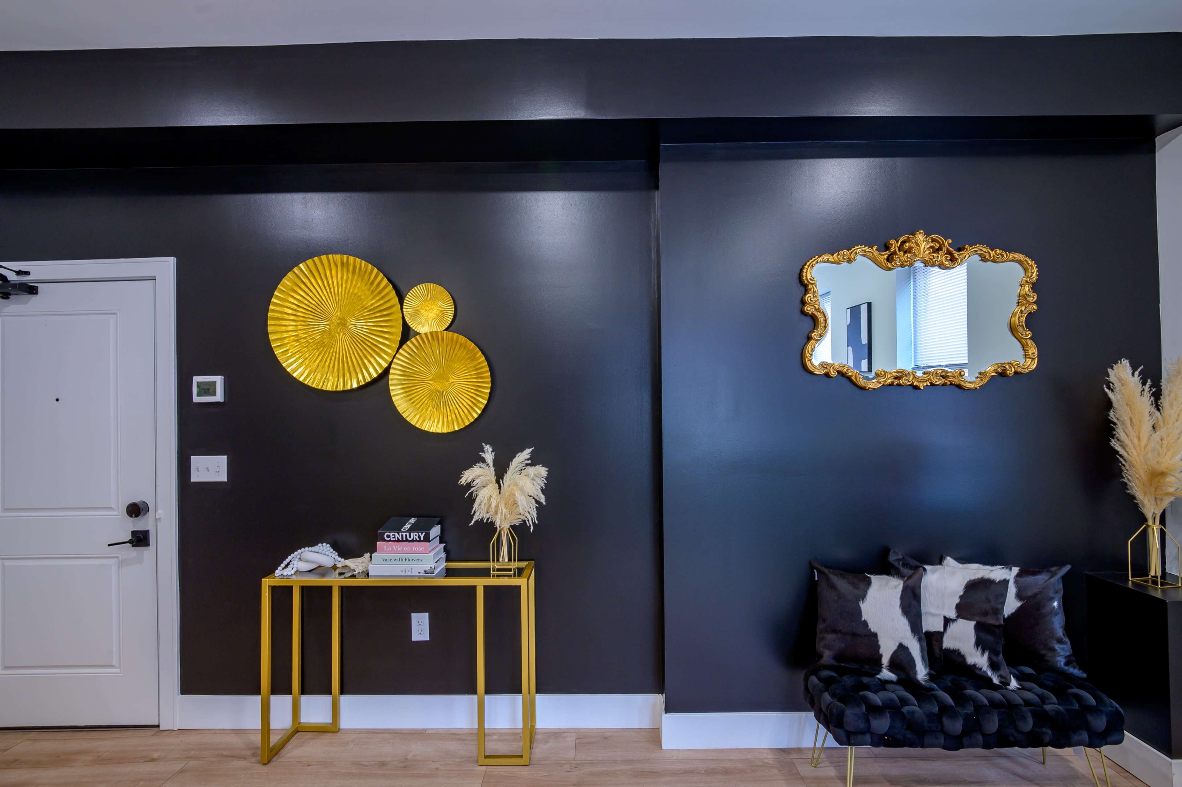 A modern entryway with a dark wall, three gold decorative circular wall pieces, a gold-framed mirror, a black and white patterned couch, and a gold side table displaying books and decorative items.