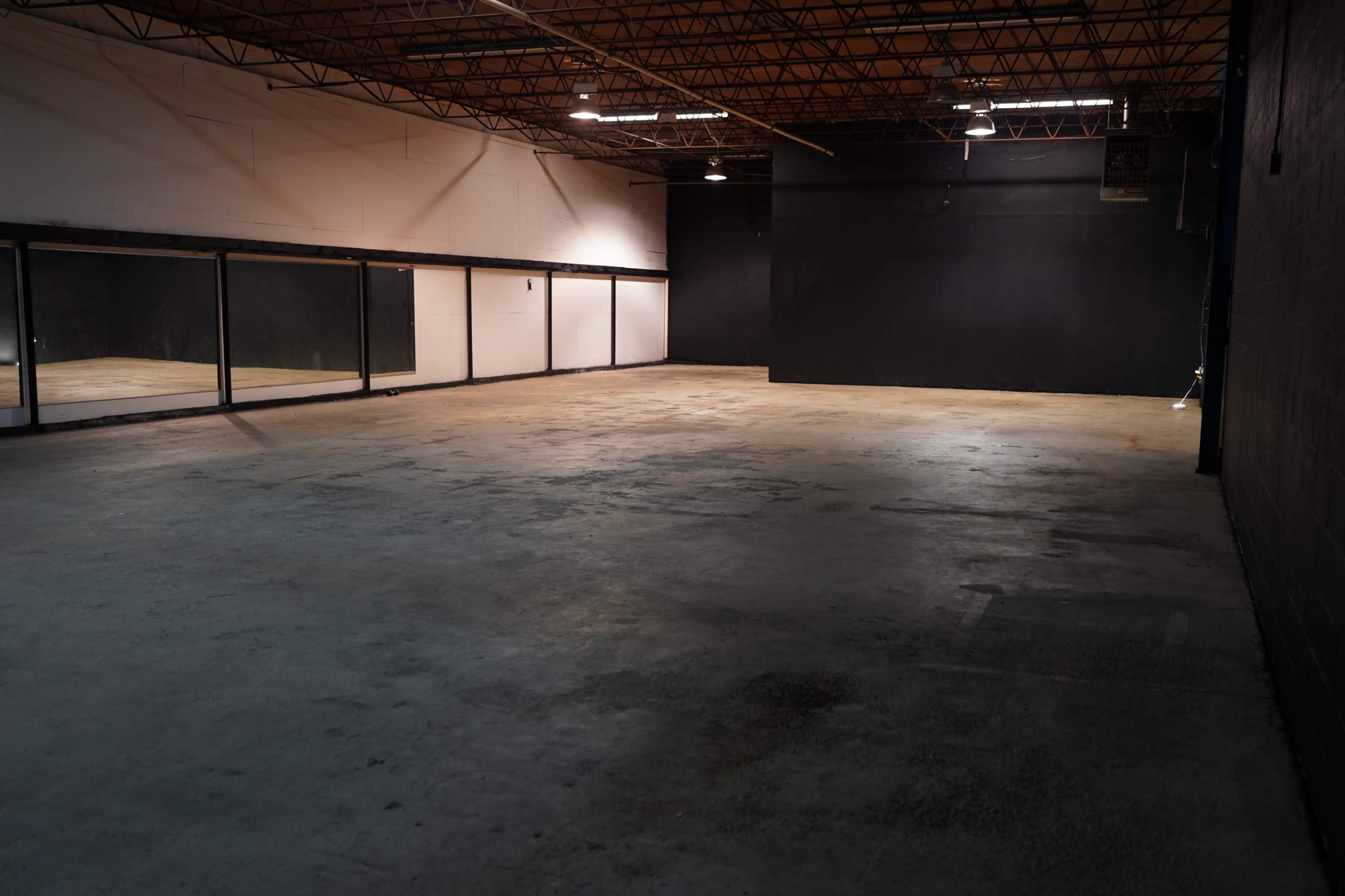 The image shows a large, empty room with concrete flooring and black walls, featuring mirrors along one side.