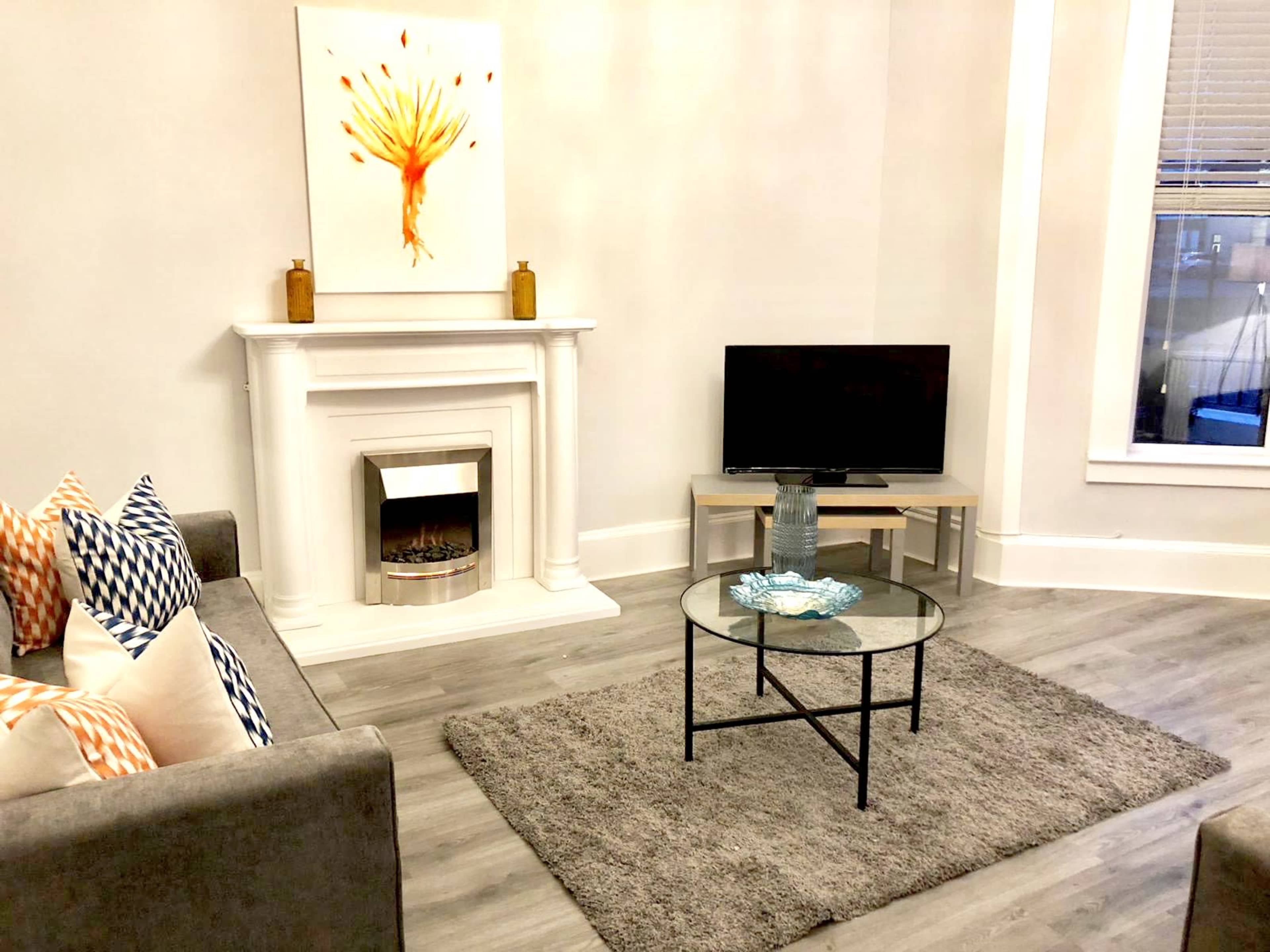 A living room features a gray sofa with patterned cushions, a round glass coffee table, a modern television, a decorative fireplace, and a large artwork on the wall.